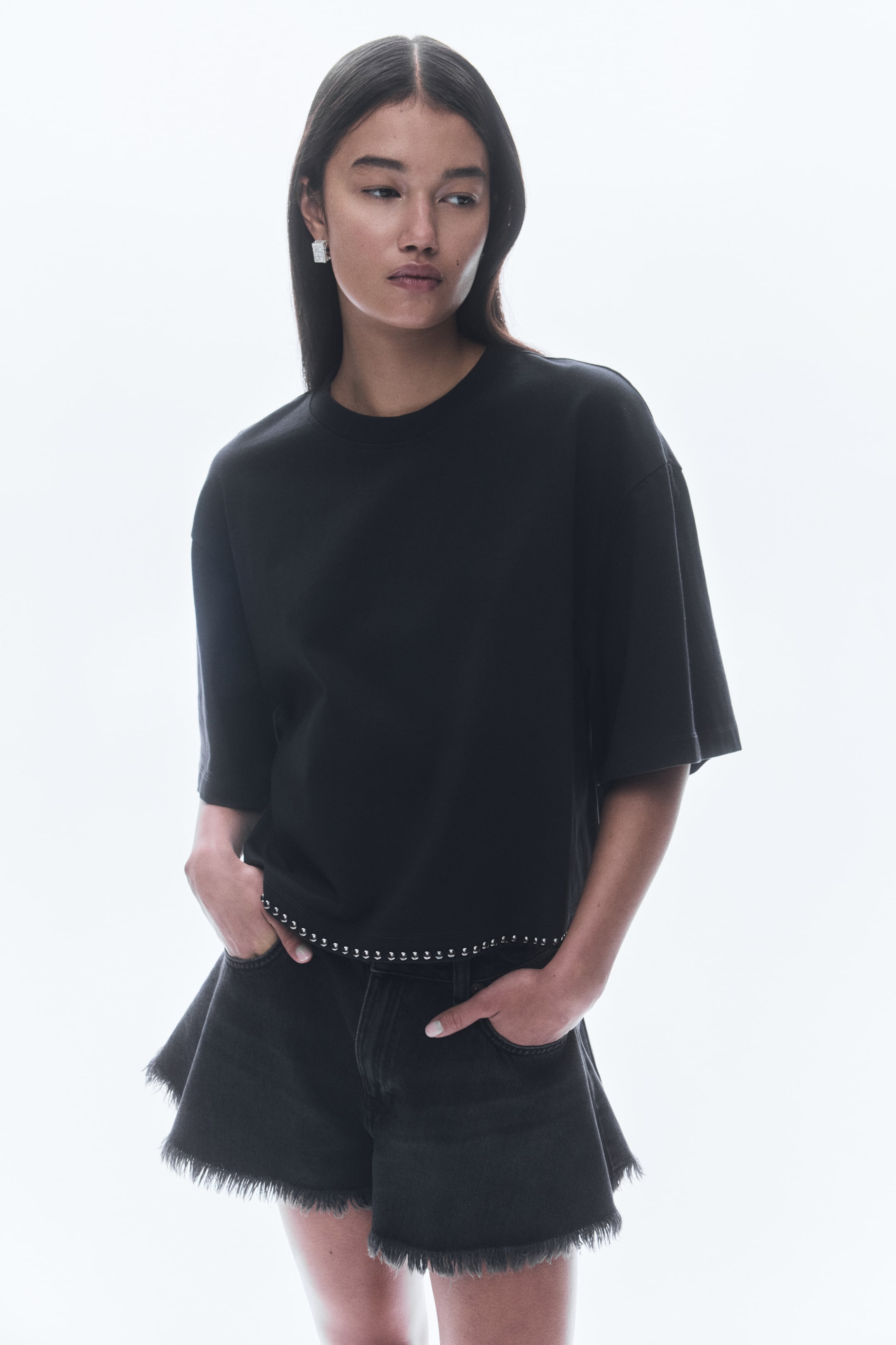 View larger image: A black crew-neck t-shirt with short, wide sleeves and a studded hemline is being presented from a slightly front-right angle. It is being styled with faded black denim shorts, which are showcasing a flared silhouette and frayed edges.