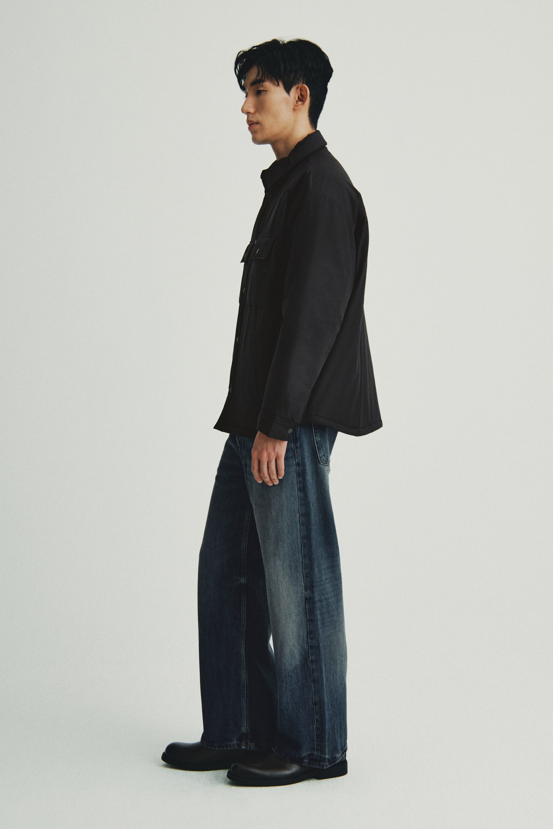 이미지 확대 보기: 검은색 깃이 달린 셔켓과 두 개의 버튼이 달린 가슴 주머니를 입은 사람이 옆모습으로 서 있으며, 넓은 다리의 블루 진이 희미한 허벅지와 어두운 둥근 앞코의 슬립온 신발과 함께 착용하고 있습니다.