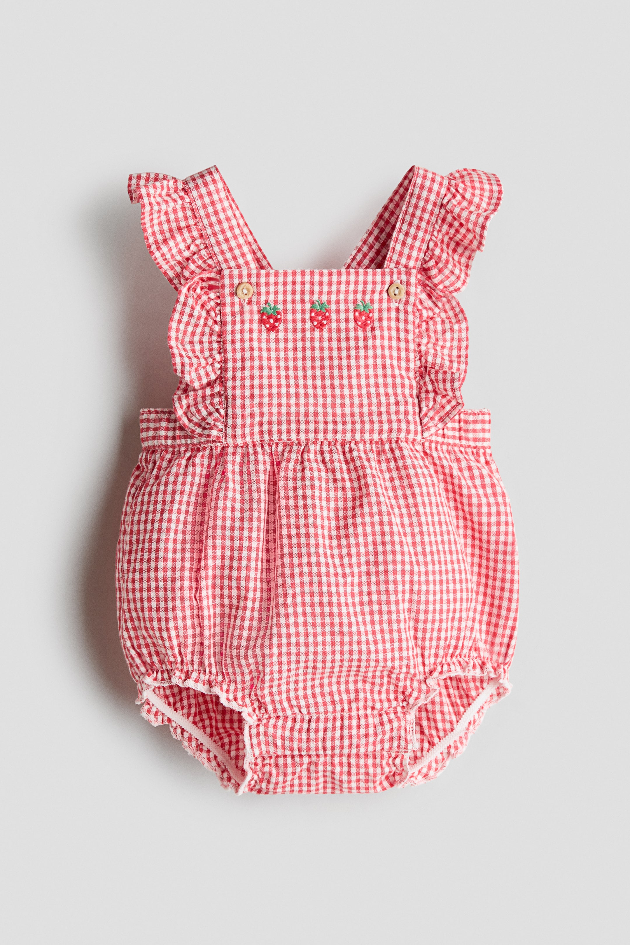 View larger image: A red and white gingham dungaree shorts is showcasing a front view, featuring frill-trimmed shoulder straps secured with wooden buttons. Three embroidered strawberries embellish the front bib, while an elasticated waist and elasticated, ruffled hems complete the design.