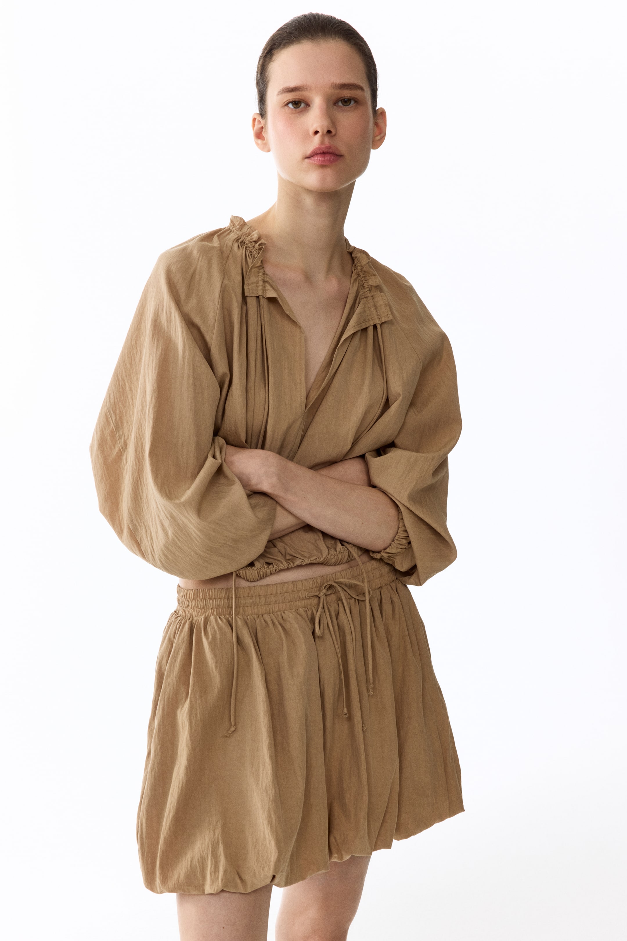 查看大圖: 這是一套棕褐色的兩件式服裝，從正面展示。上衣具有聚攏的領口、豐盈的長袖並配有彈性袖口，還有一個皺褶彈性腰身。搭配的短褲則展示了彈性抽繩腰部和聚攏的燈籠褲腿，完整了這套服裝的造型。