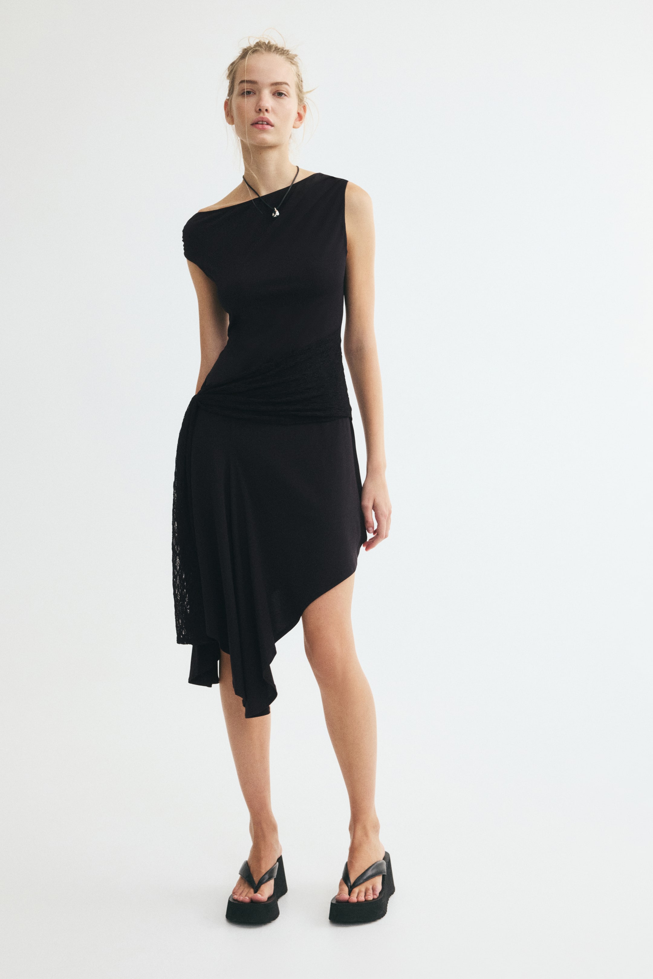 View larger image: The model is wearing a black draped lace-panel dress featuring an asymmetrical neckline, a ruched waist with a lace panel, and an asymmetrical draped skirt. The dress is styled with a black cord pendant necklace and black platform thong sandals.