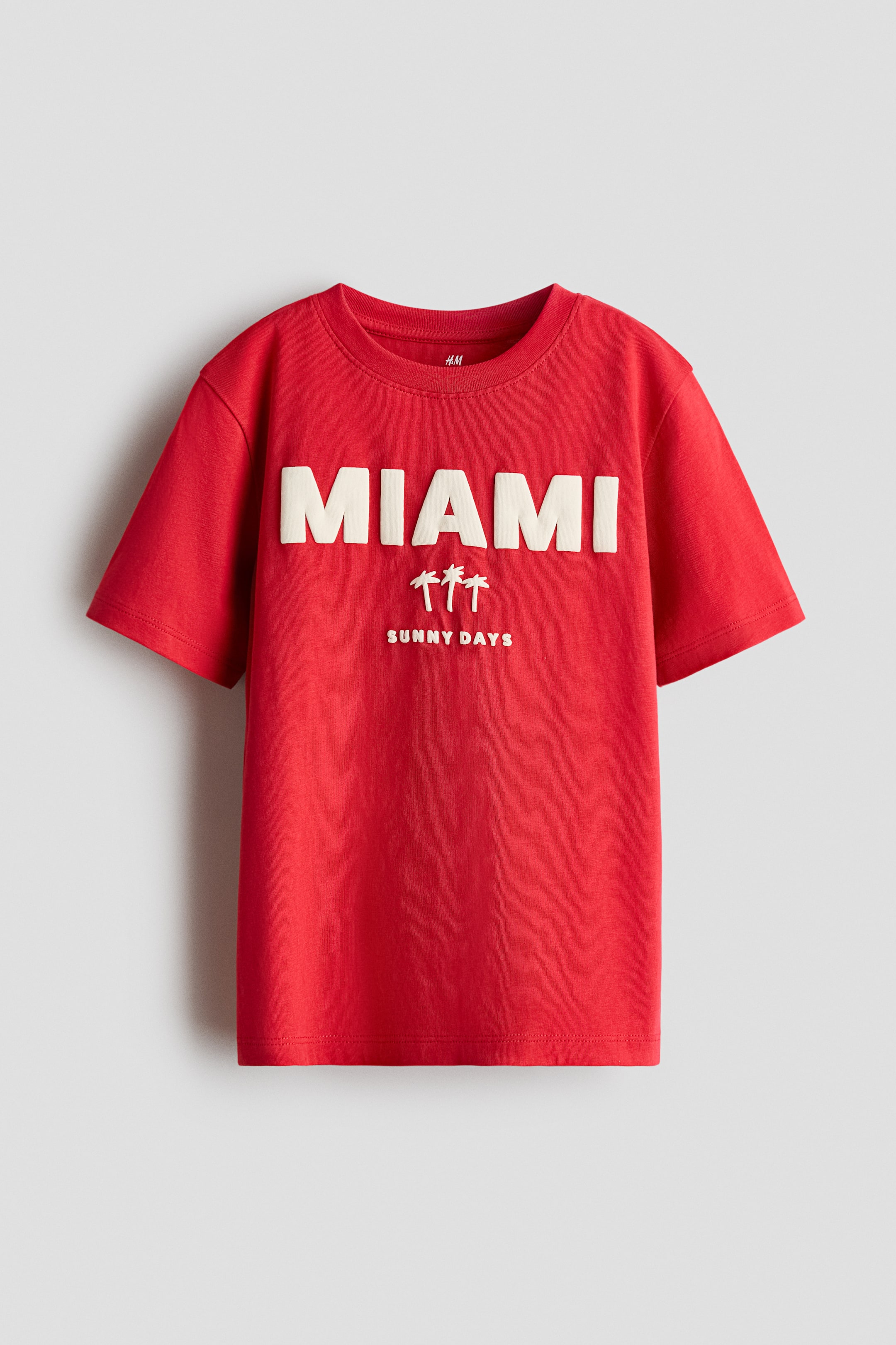 View larger image: A red T-shirt in soft cotton jersey is displayed from the front, featuring a round, rib-trimmed neckline and gently dropped shoulders. It showcases a light-coloured print with "MIAMI" in raised letters, and three palm trees with "SUNNY DAYS" text below.