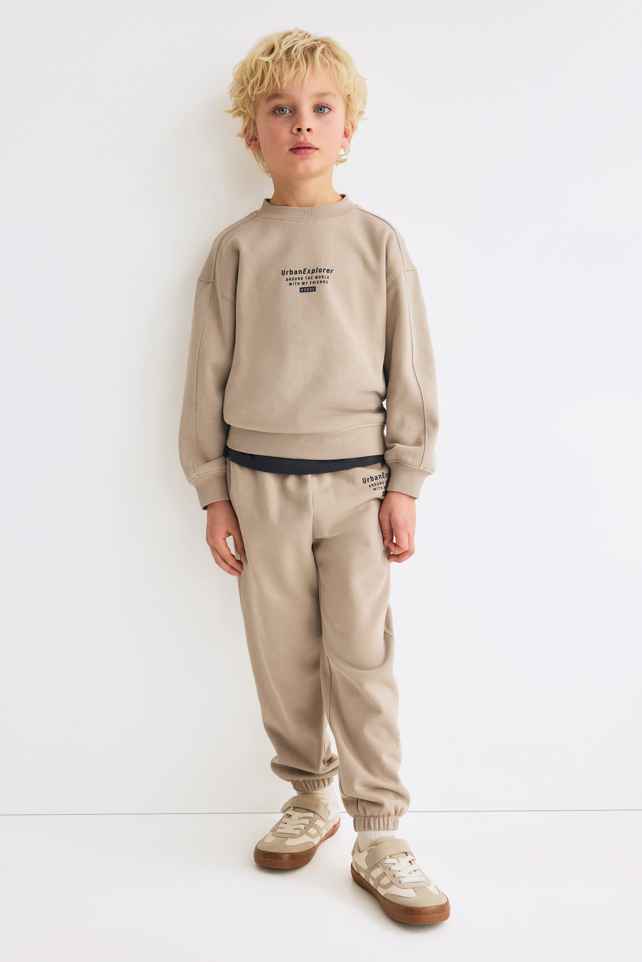 Grösseres Bild ansehen: Ein beiges Sweatshirt- und Jogger-Set wird von vorne präsentiert. Das Sweatshirt verfügt über einen runden, mit Rippbündchen versehenen Ausschnitt, überschnittene Schultern, lange Ärmel mit dekorativen Nähten, gerippte Bündchen und Saum sowie den Schriftzug "UrbanExplorer" auf der Brust. Die passenden Jogger haben einen elastischen Bund, Seitentaschen, verdeckte Elastik an den Säumen und den "UrbanExplorer"-Schriftzug auf dem linken Oberschenkel. Unter dem Sweatshirt ist ein schwarzes Unterhemd sichtbar.