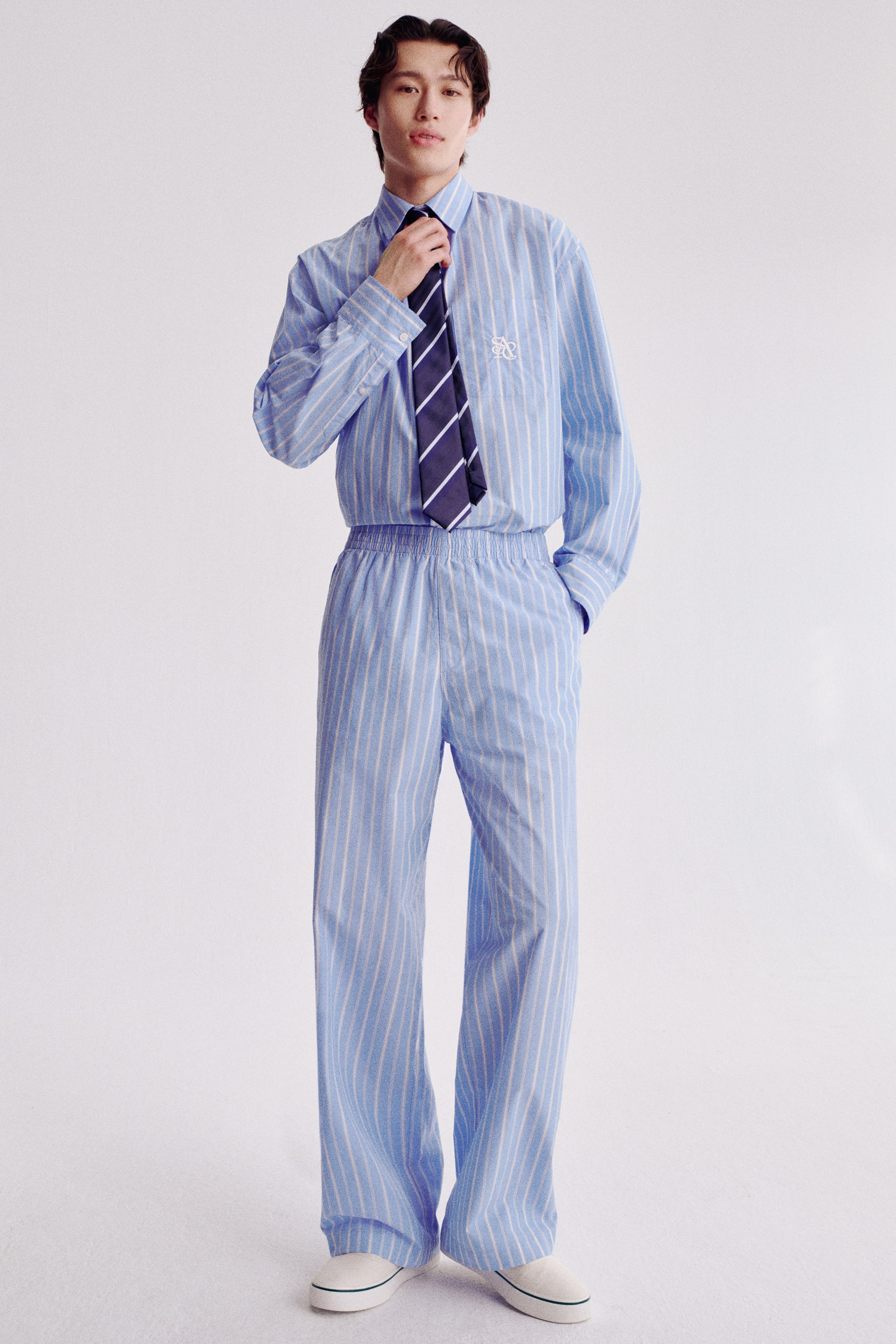 Größeres Bild ansehen: Ein hellblau-weiß gestreiftes Button-Down-Hemd und dazu passende weite Hosen werden von vorne präsentiert. Das Hemd mit Kragen, langen Ärmeln und einem weißen, gestickten Logo auf der linken Brust wird mit einer marineblau-weiß gestreiften Krawatte gestylt, die am Hals angepasst wird; die Hosen haben einen elastischen Bund.