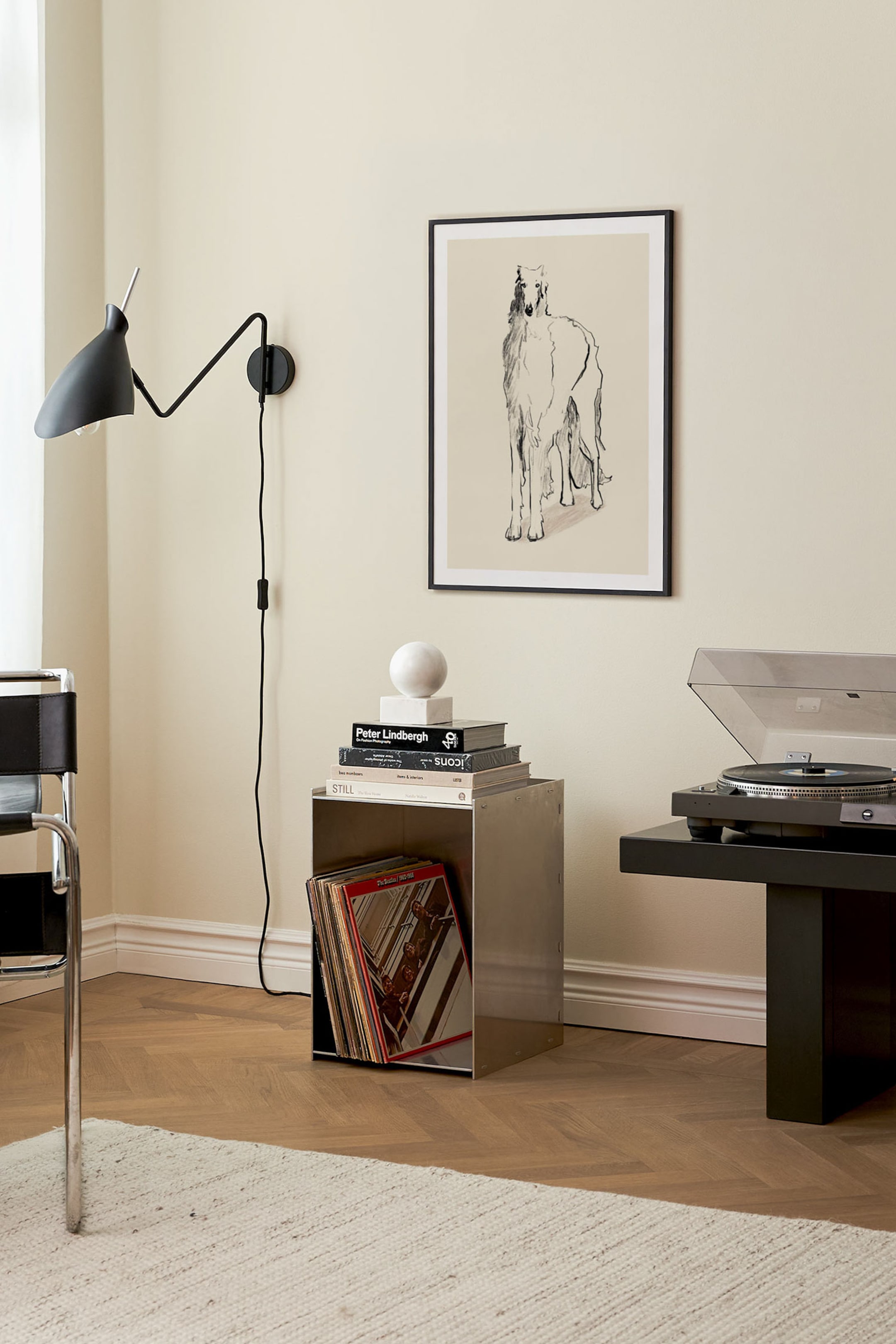 Se større billede: Et kubisk sidebord i børstet rustfrit stål står i en vinkel og fremhæver sin metalliske konstruktion og åbne frontkompartment. Det åbne rum rummer en række vinylplader, mens flere bøger og en sfærisk stenskulptur ligger på den flade topflade.