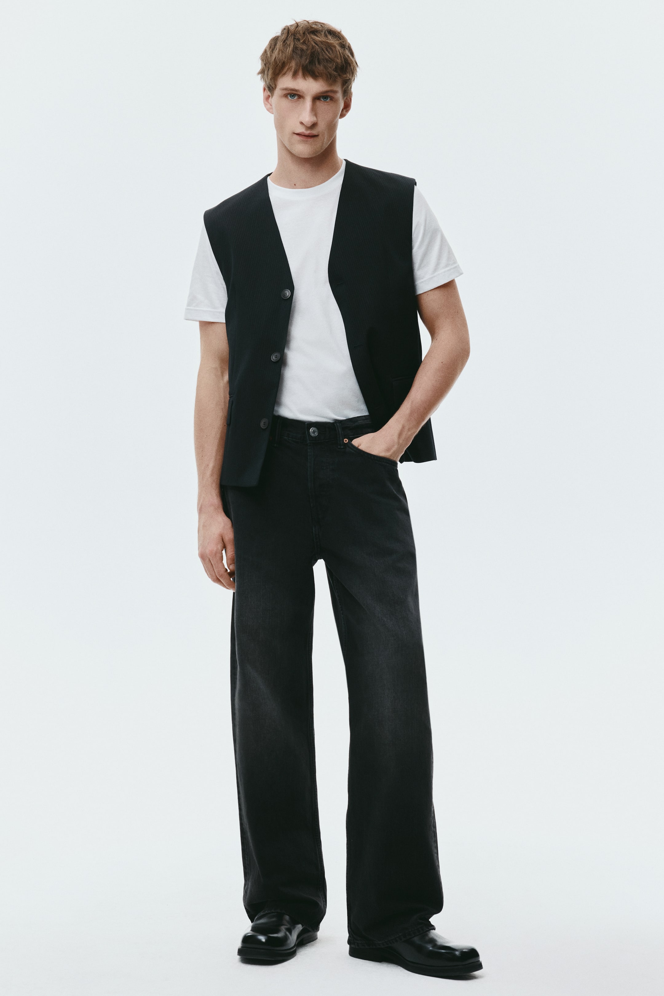 Näytä suurempi kuva: Tyylitelty kokonaisuus sisältää mustan V-kaula-aukkoinen liivin, jossa on kolme tummaa nappia ja hienovarainen pystysuuntainen tekstuuri, kerrostettuna valkoisen pyöreäkauluksisen t-paidan päälle. Sen kanssa on yhdistetty tummanpesuiset, leveälahkeiset mustat farkut ja kiillotetut mustat nahkaloaferit. Kokonaisuus on esitetty edestäpäin.