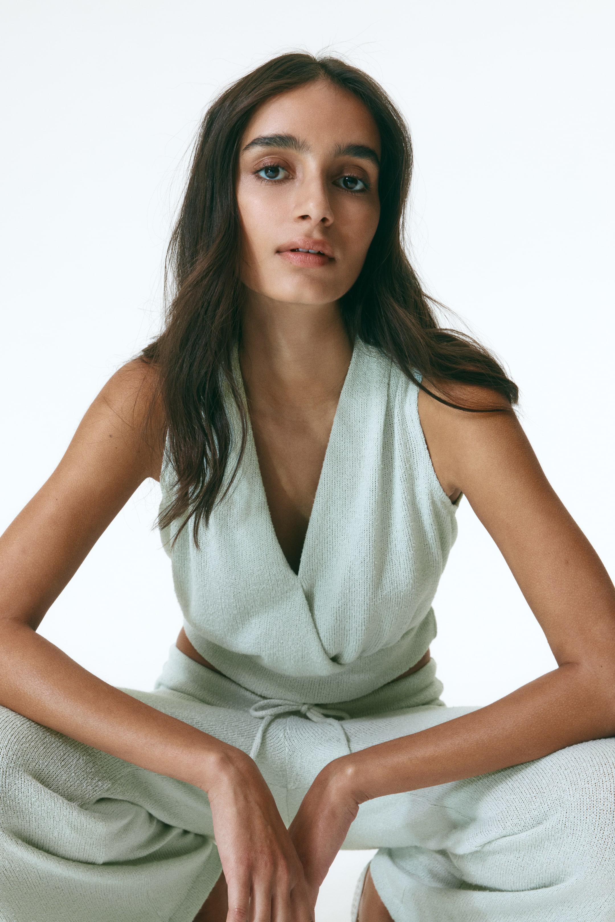 View larger image: The model is showcasing a short, light green knitted top featuring a deep V-neckline and a sewn-in wrapover front, paired with a sleeveless design. She is also wearing matching knitted trousers.