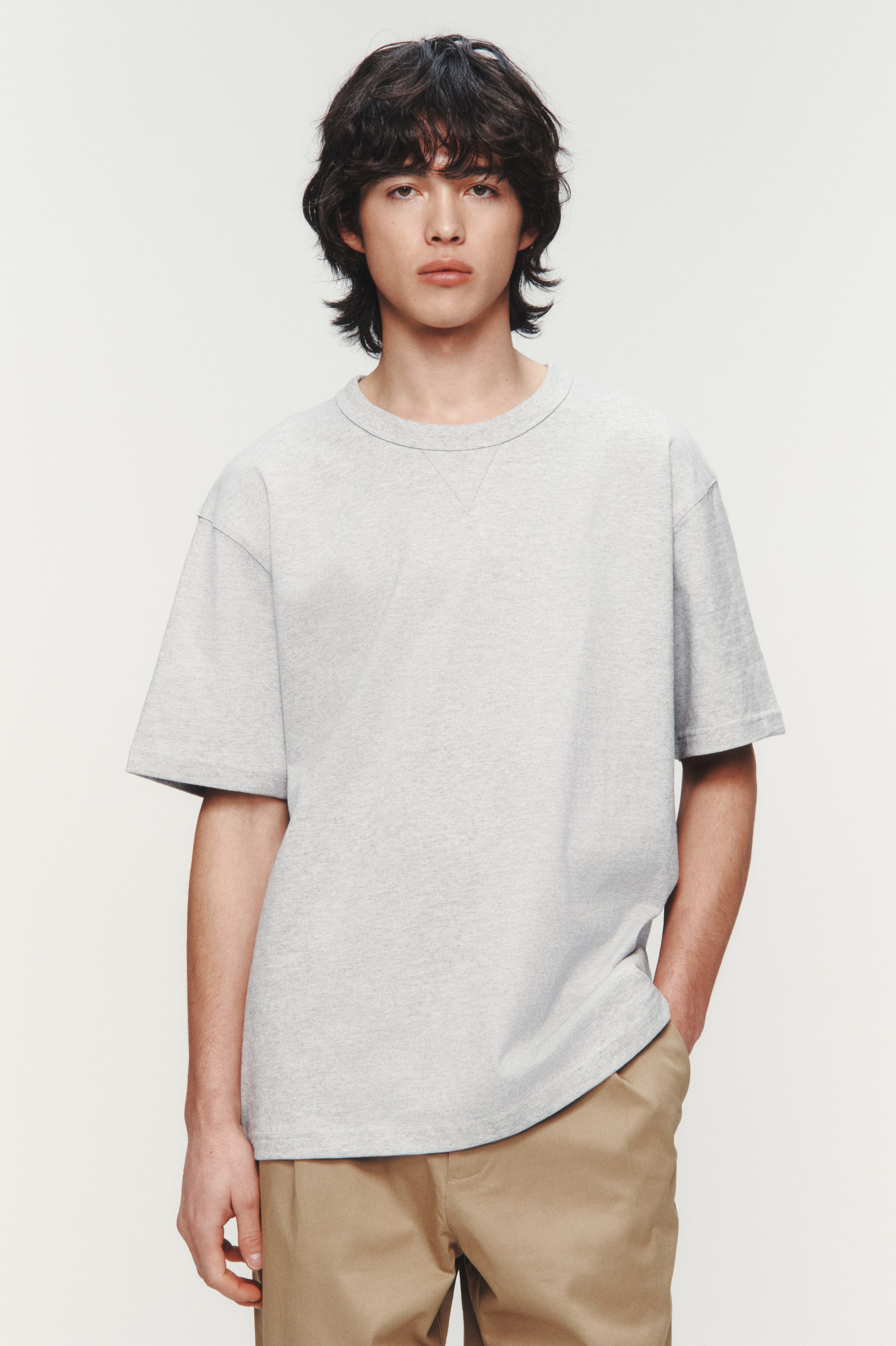 拡大画像を表示: ライトグレーのクルーネックTシャツを着用しており、前面を向いています。リラックスフィット、ショートスリーブ、ネックライン下のVステッチディテールが特徴です。