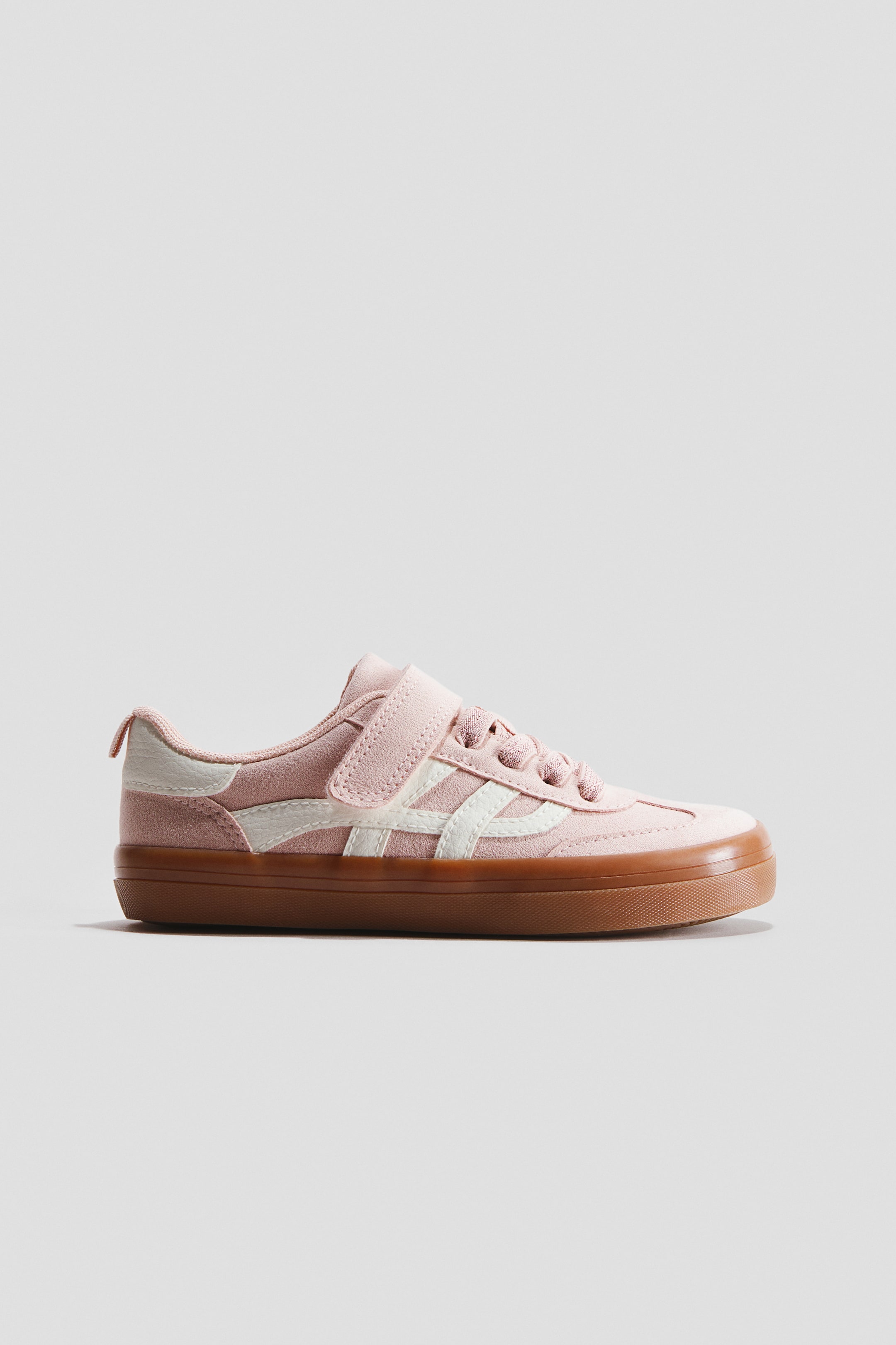 Zobacz większy obraz: Różowy sneaker prezentuje widok z boku, posiadający cholewkę przypominającą zamsz z różowymi sznurówkami, pojedynczym paskiem na rzep oraz białymi, kanciastymi paskami po bokach. Dodatkowo, widoczne są biała zakładka na pięcie, brązowa gumowa podeszwa oraz wzmocniony, zaokrąglony nosek.