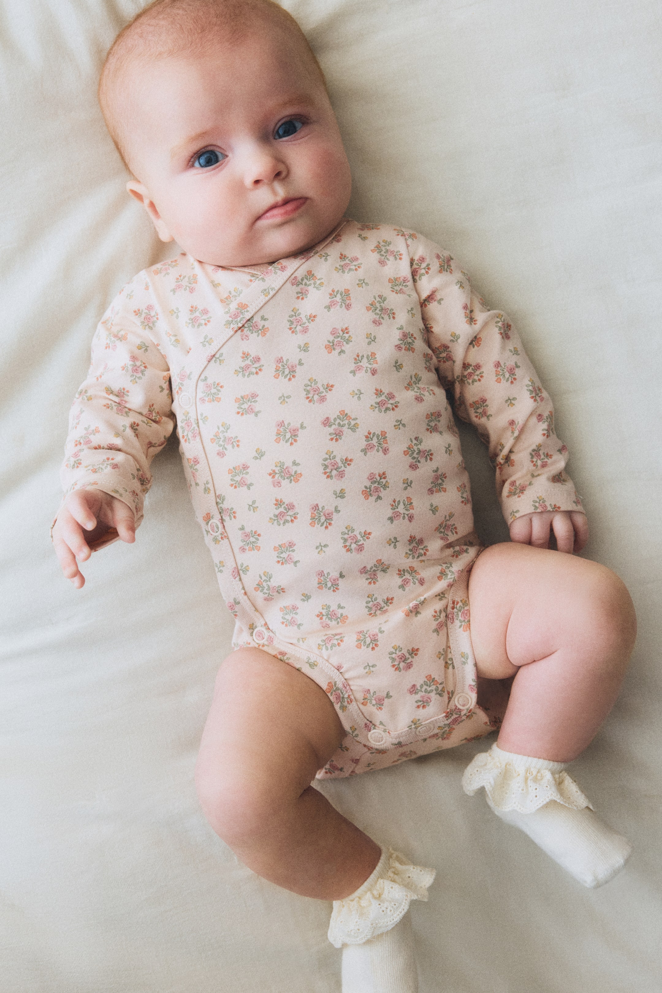 Grösseres Bild ansehen: Ein hellrosa Baby-Strampler wird aus der Vogelperspektive präsentiert, mit langen Ärmeln und einem kleinen Blumenmuster mit rosa, grünen und orangefarbenen Blumen. Er ist im Kimono-Stil mit einer Wickelvorderseite gestaltet, die mit Druckknöpfen an der Seite und zusätzlichen Druckknöpfen im Schritt gesichert wird.