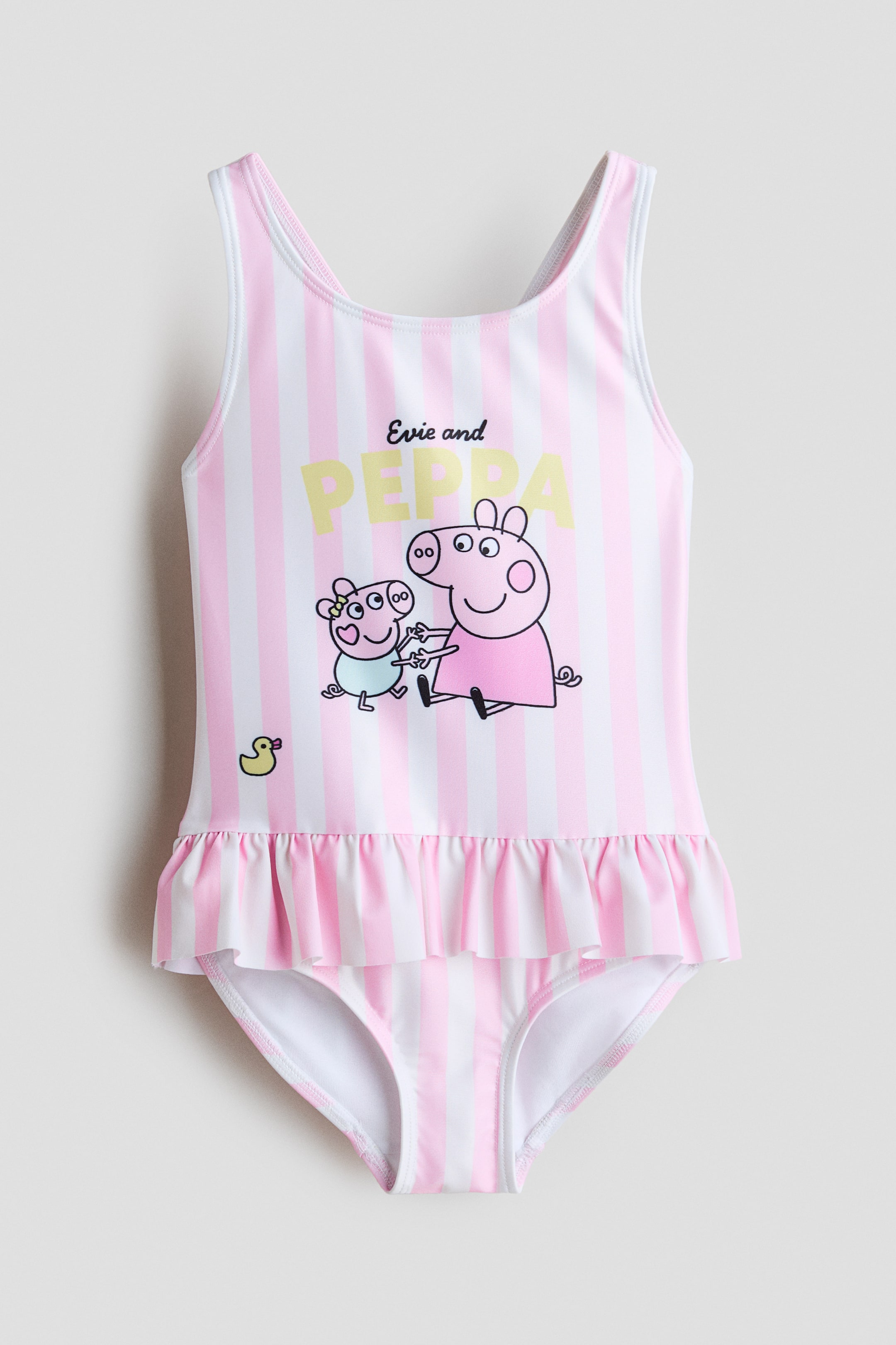 Agrandir l'image: Un maillot de bain imprimé présente des rayures verticales blanches et roses avec un graphisme de deux personnages de dessin animé en forme de cochon, un petit canard jaune, et le texte "Evie and Peppa" sur le devant. Il est conçu avec de larges bretelles qui se croisent dans le dos et un volant avec des fronces à la taille.
