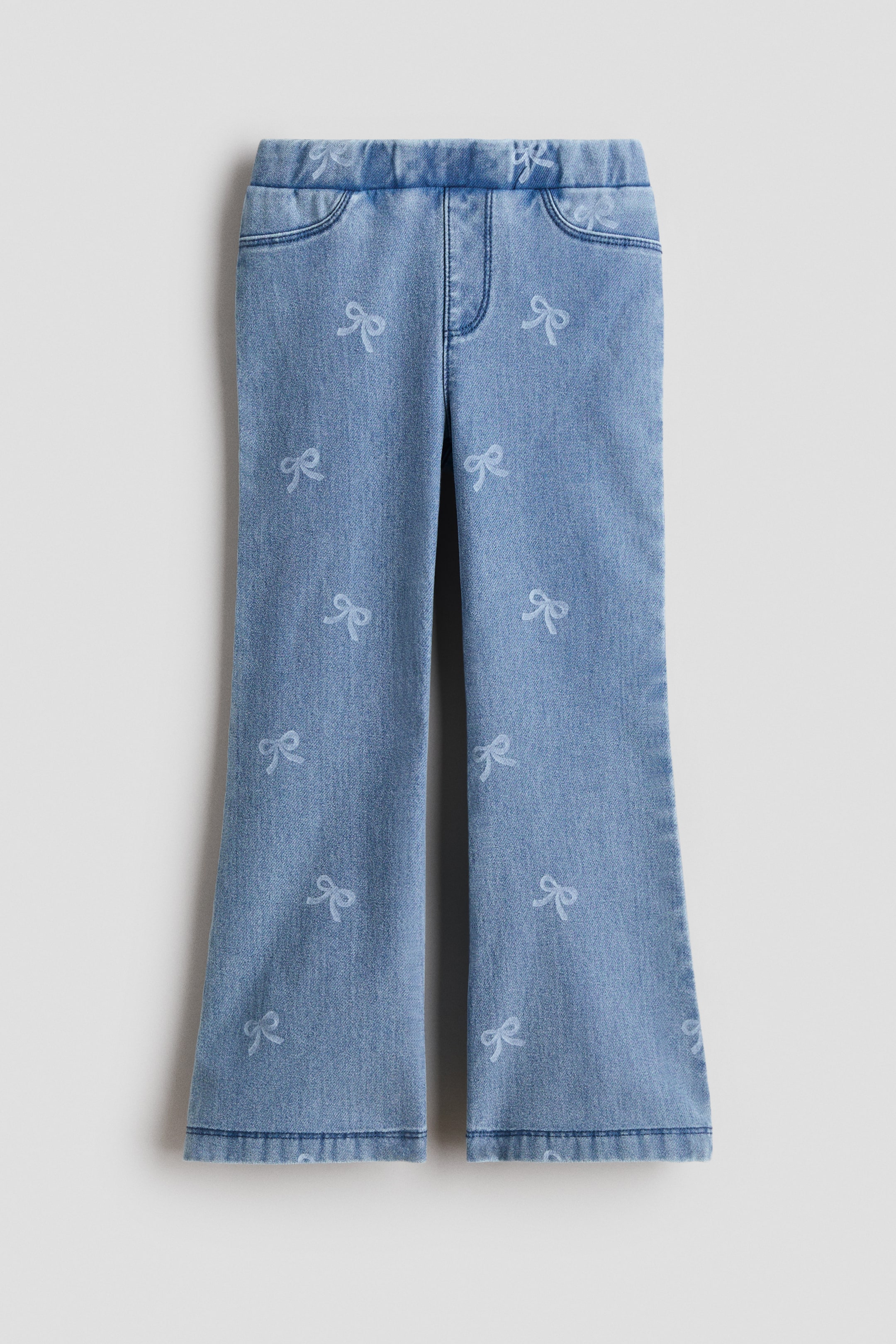 Ampliar la imagen: Los jeggings se muestran de frente en un jersey de aspecto denim azul claro, con piernas acampanadas y un motivo de lazos en toda la prenda. Presentan cintura elástica, bragueta falsa y bolsillos frontales decorativos.