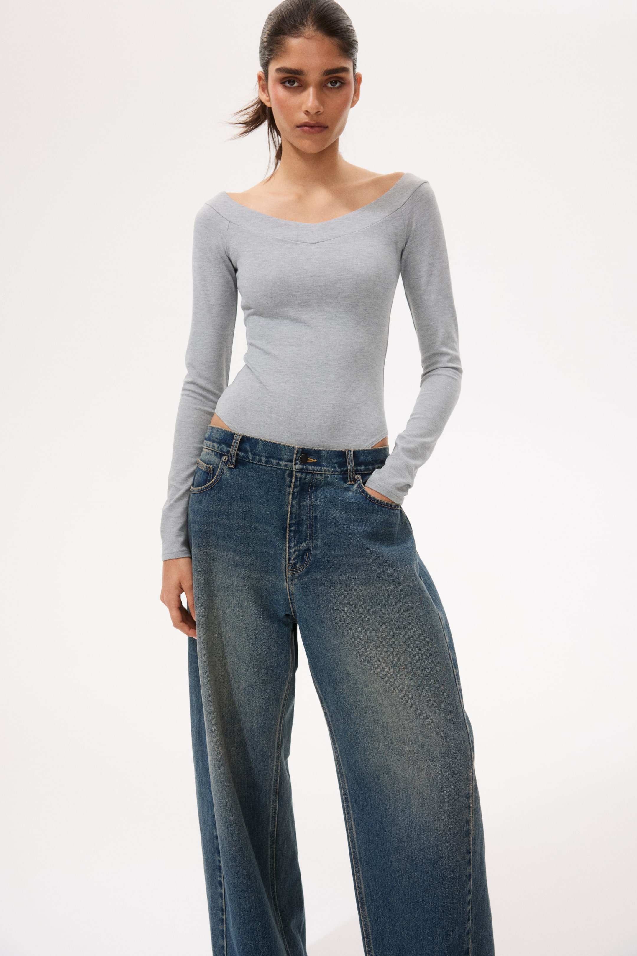 View larger image: A grey, long-sleeved bodysuit featuring an off-the-shoulder V-neckline. It fits snugly and is styled tucked into wide-leg blue jeans, highlighting its high-cut design.