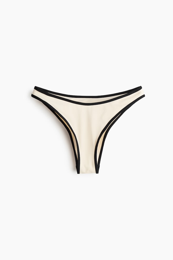 A cream thong-style brief lies flat, showing the front with black trim outlining the waistband and high-cut leg openings. - 1
