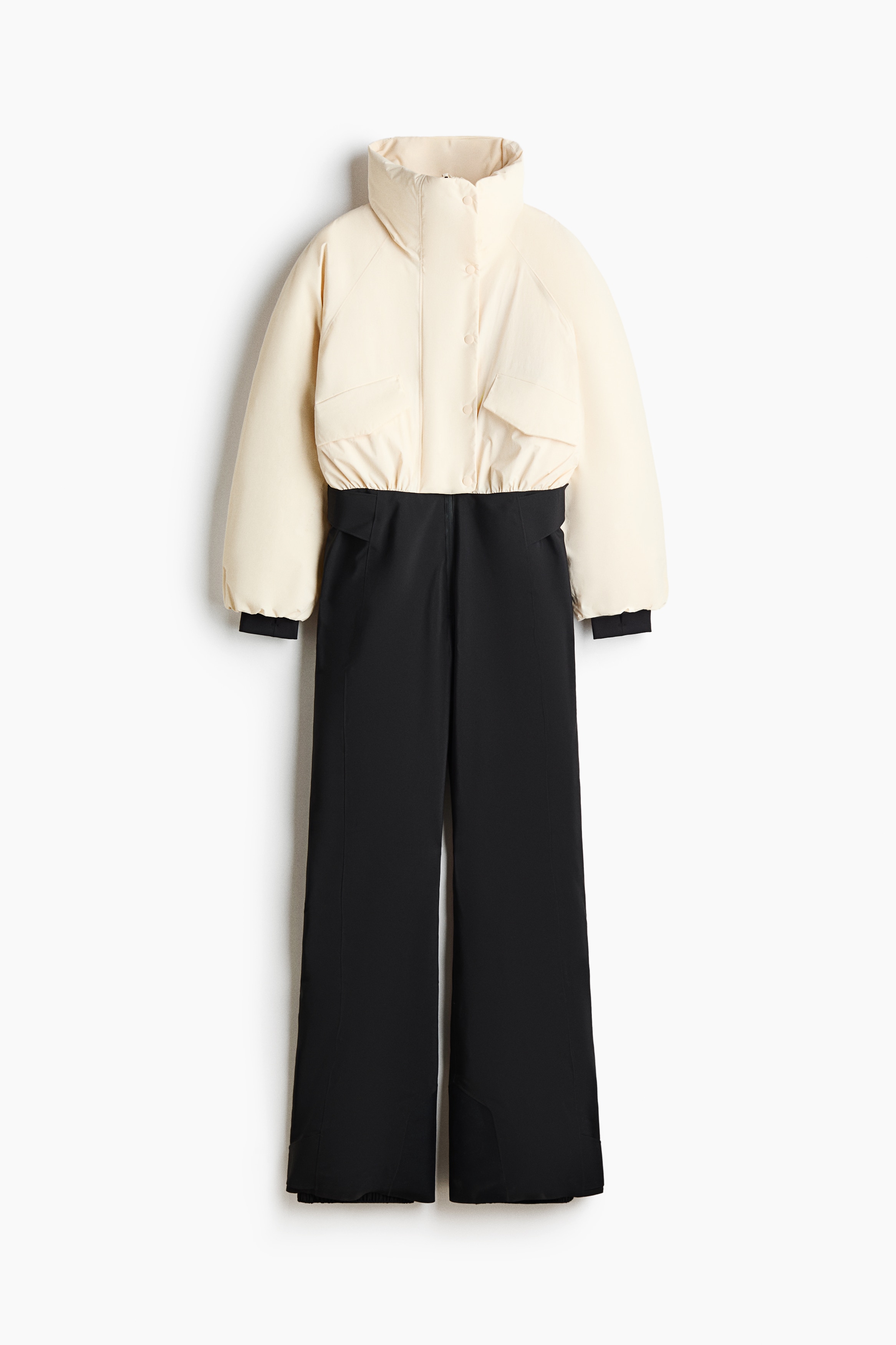 Une doudoune courte blanc cassÃ© dotÃ©e dâun col montant structurÃ© est portÃ©e par le mannequin, qui regarde vers le bas de profil. La veste prÃ©sente des manches volumineuses avec des poignets Ã©lastiquÃ©s foncÃ©s et est associÃ©e Ã  un pantalon de ski noir taille haute ainsi quâÃ  des bottes de ski noires.