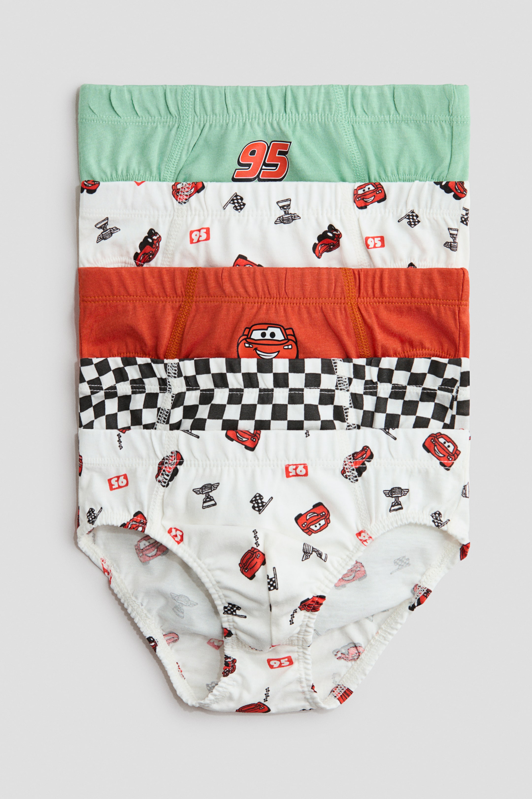 Voir une image plus grande: Un ensemble de 5 boxers pour garçons est présenté de face, empilés verticalement. Chaque boxer est doté d'une ceinture élastique et d'ouvertures pour les jambes, avec divers motifs inspirés de "Cars" incluant des voitures rouges, des drapeaux à damier, et le numéro 95, apparaissant sur des tissus vert clair, blanc, orange, et à damier noir et blanc.
