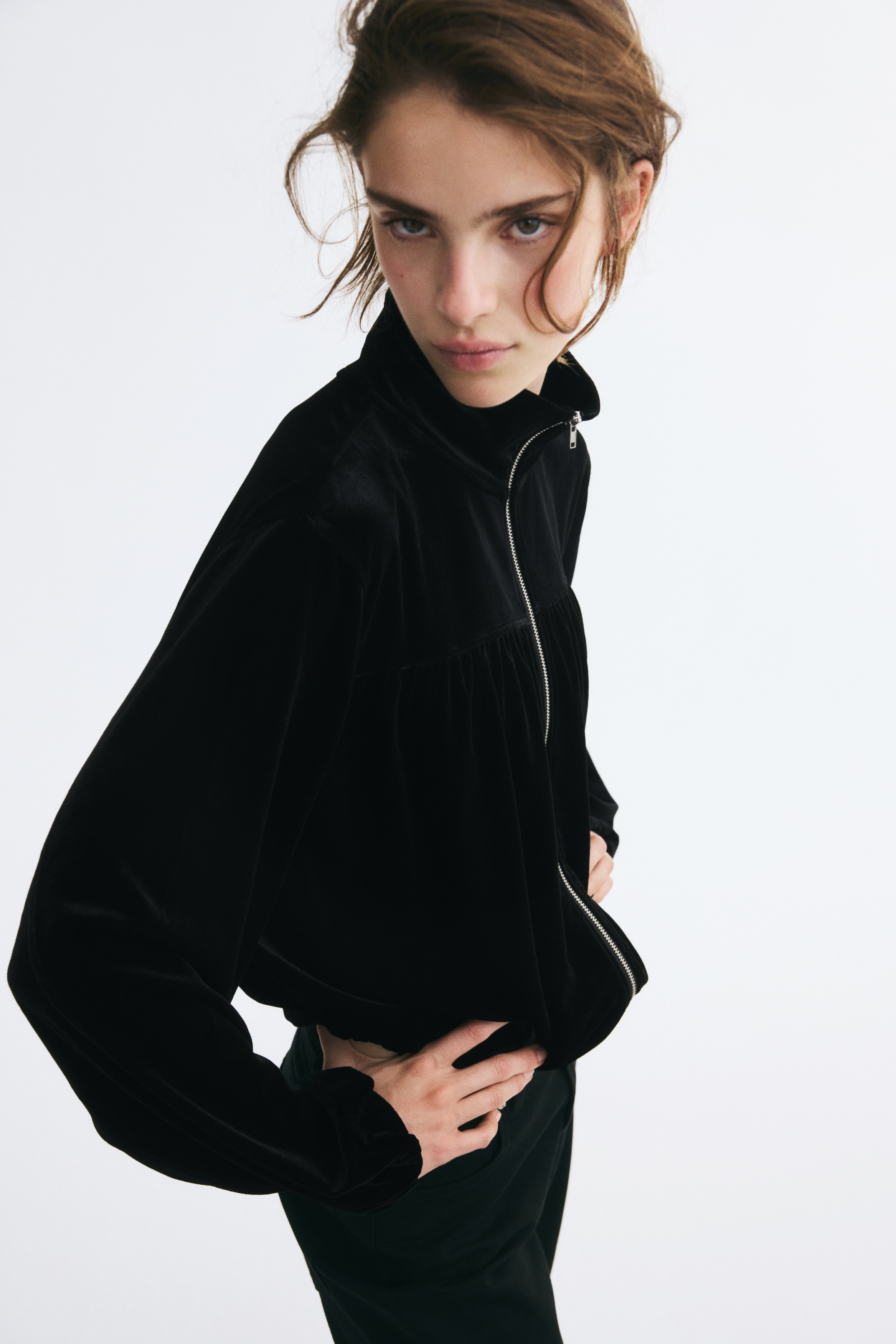 A black velvet jacket is shown from the front, featuring a high mock neck and a full-length silver zip down the centre. The design features a horizontal chest yoke with gathered fabric draping down to an elasticated hem, accompanied by long sleeves with elasticated cuffs.