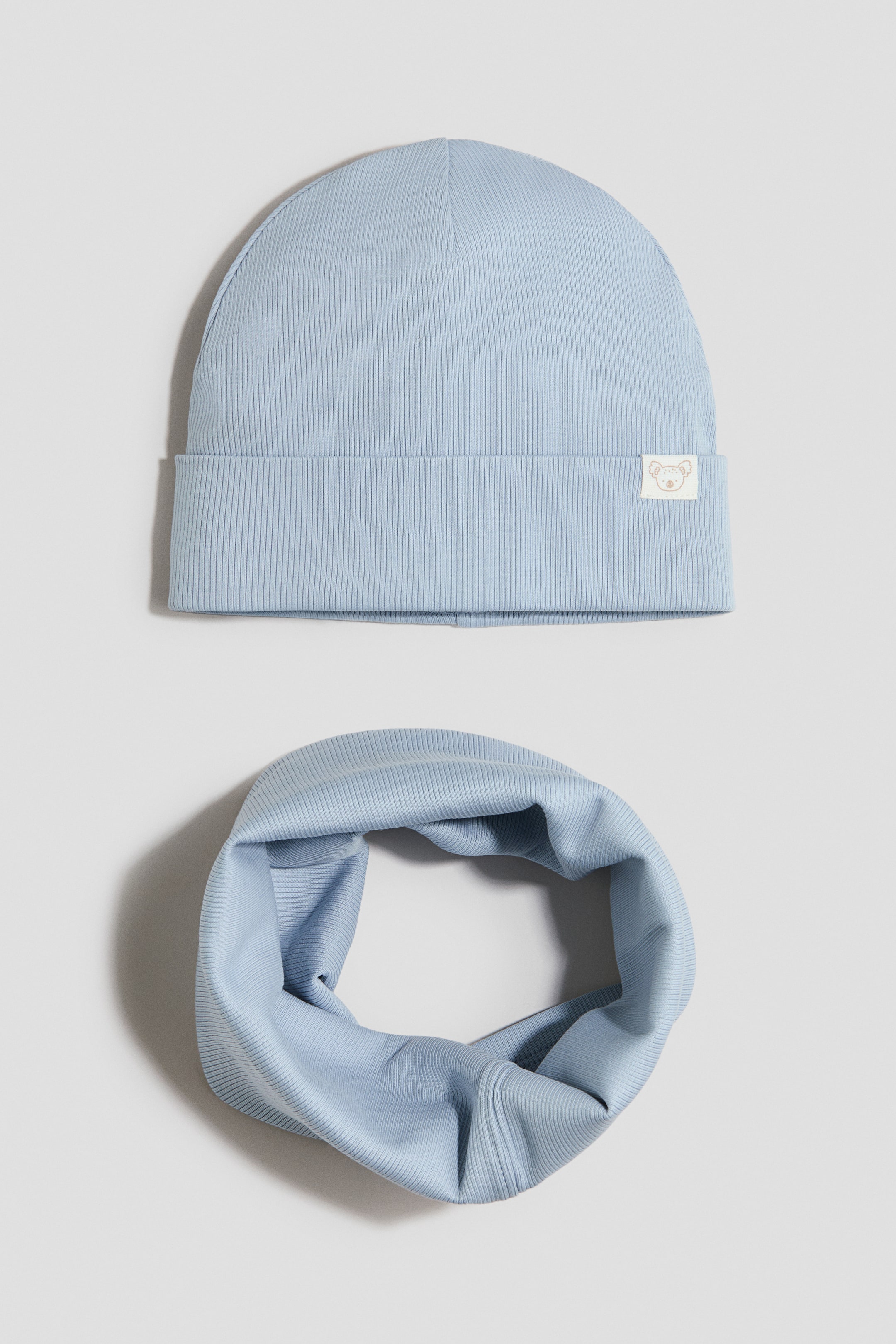 View larger image: A 2-piece jersey set features light-blue items, including a ribbed beanie and a matching ribbed neck warmer. The beanie faces forward, revealing a turned-up brim with a small white label bearing an animal illustration, while the neck warmer is arranged in a circular, folded shape.