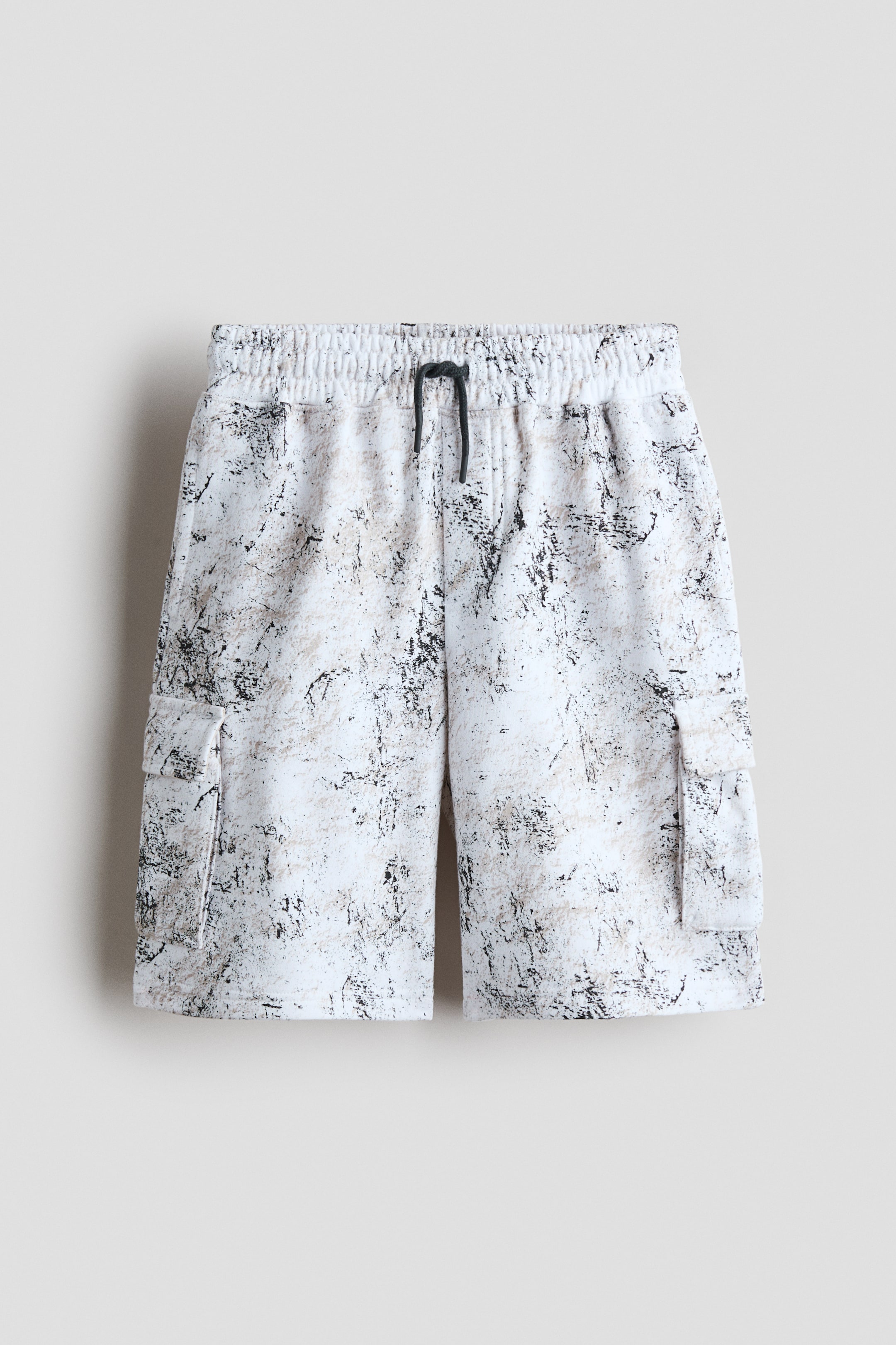 Zobacz większy obraz: Szorty charakteryzują się białą bazą z abstrakcyjnym czarno-beżowym wzorem, elastycznym pasem z czarnym sznurkiem oraz dwoma kieszeniami cargo z klapami po bokach.