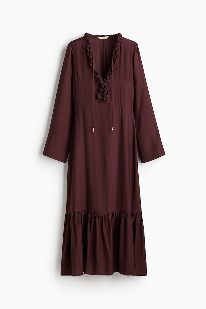 A dark red maxi dress in woven fabric is displaying its FRONT. It is featuring a deep, ruffle-trimmed V-neckline with spaghetti ties that are ending in gold-toned beads. Long sleeves with wide cuffs are extending from the bodice, and an angular seam is defining the waist of a flared skirt that is showcasing a gathered tier above the hem. - 1
