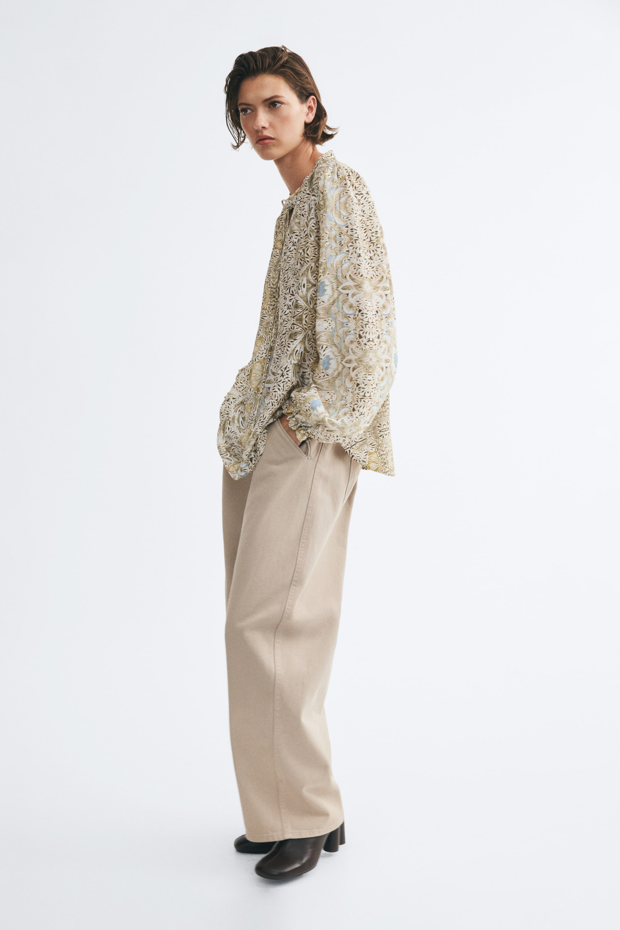 View larger image: A loose-fit blouse with an intricate beige and blue pattern is being worn by the model, featuring long sleeves with elasticated, frilled cuffs. The blouse is paired with beige wide-leg trousers and dark boots, with the model standing in a side-profile pose.