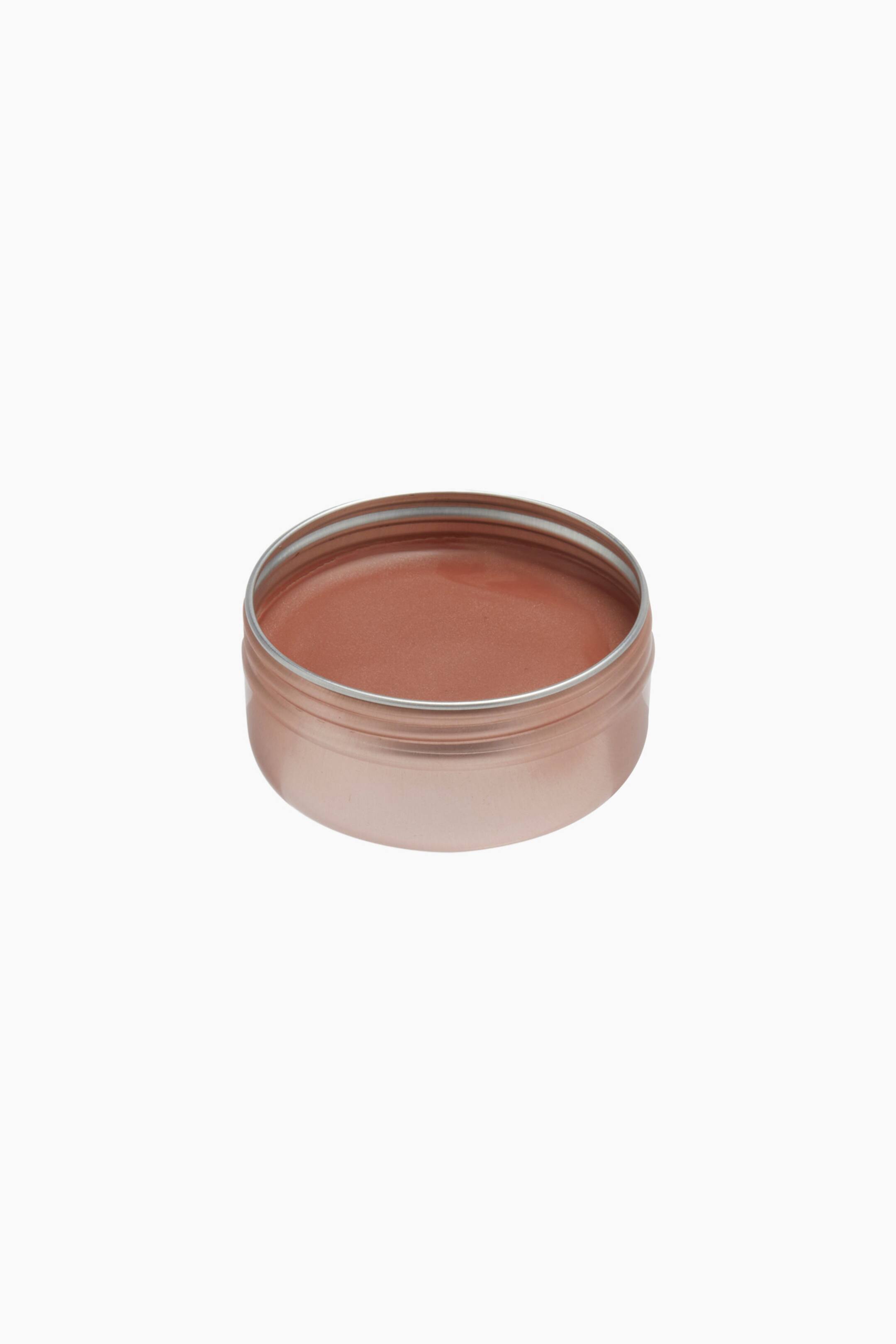 Größeres Bild ansehen: Eine runde Metalldose in Roségold enthält einen glatten Creme-Balsam in Terrakotta-Farbe und ist aus einer leicht erhöhten Dreiviertelansicht zu sehen, wodurch die glänzende, feste Oberfläche zur Geltung kommt.