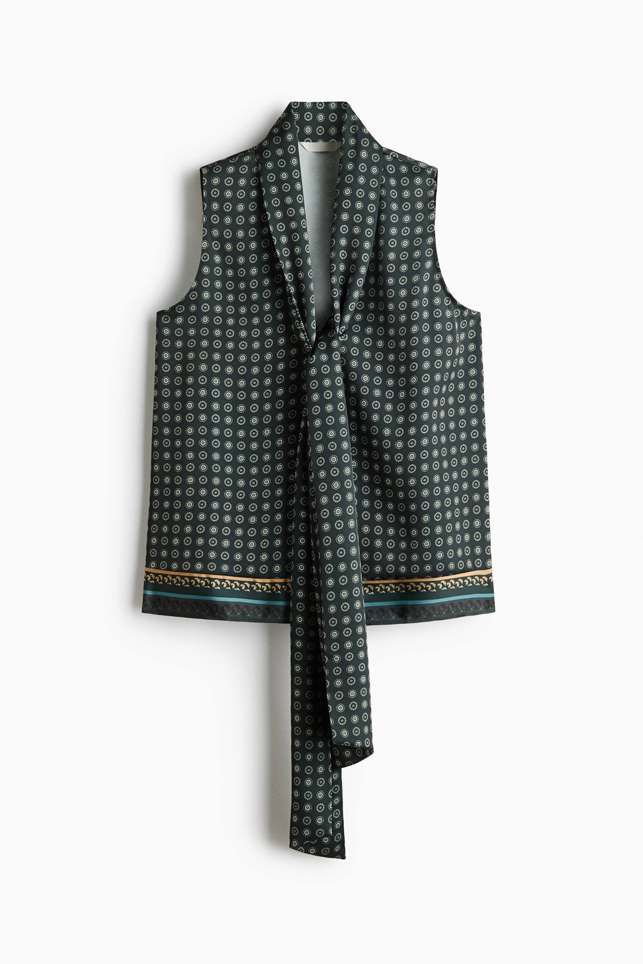View larger image: A front-facing sleeveless blouse features an all-over small circular geometric pattern on a dark green base. The top has a V-neckline with long, draped neckties hanging down the front and is finished with a decorative gold and teal border print along the straight hem.