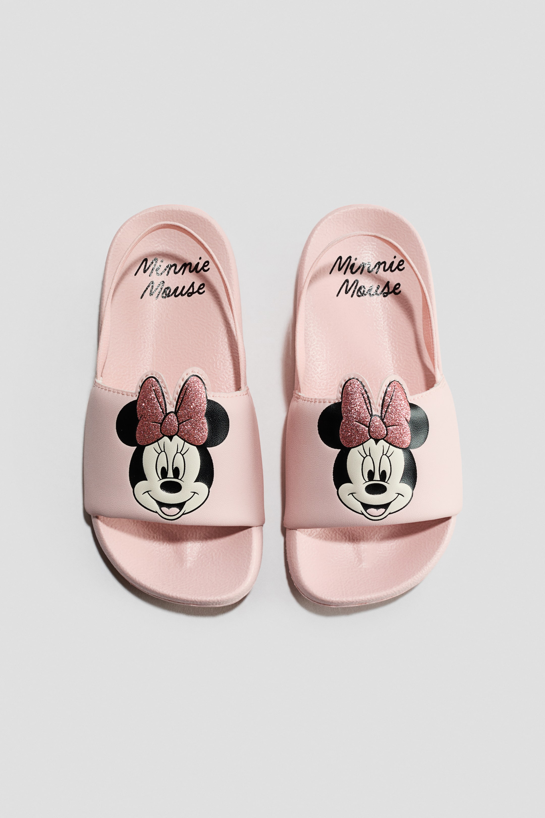 View larger image: A pair of light pink pool shoes is facing front, each boasting a wide front strap adorned with a Minnie Mouse character face and a glittery pink bow. An elasticated heel strap secures the back of each shoe, with "Minnie Mouse" text visible on the moulded footbeds.