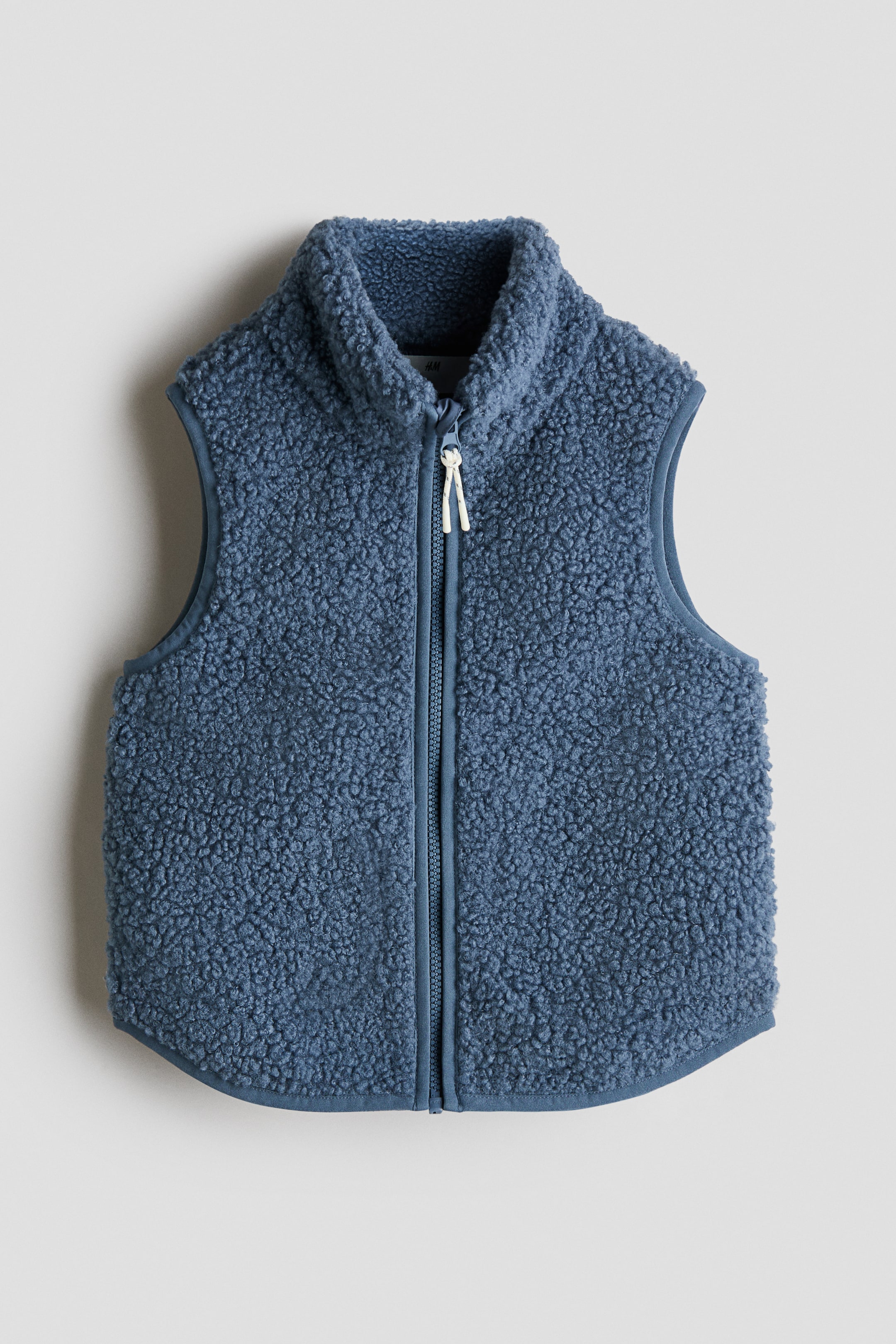 Grotere afbeelding bekijken: Een blauwe boucl� fleece bodywarmer wordt frontaal gepresenteerd, met een opstaande kraag, een volledige ritssluiting aan de voorkant met twee witte koordritstrekkers en een mouwloos ontwerp. De armsgaten en de gebogen zoom aan de onderkant zijn afgewerkt met bijpassende blauwe biezen.