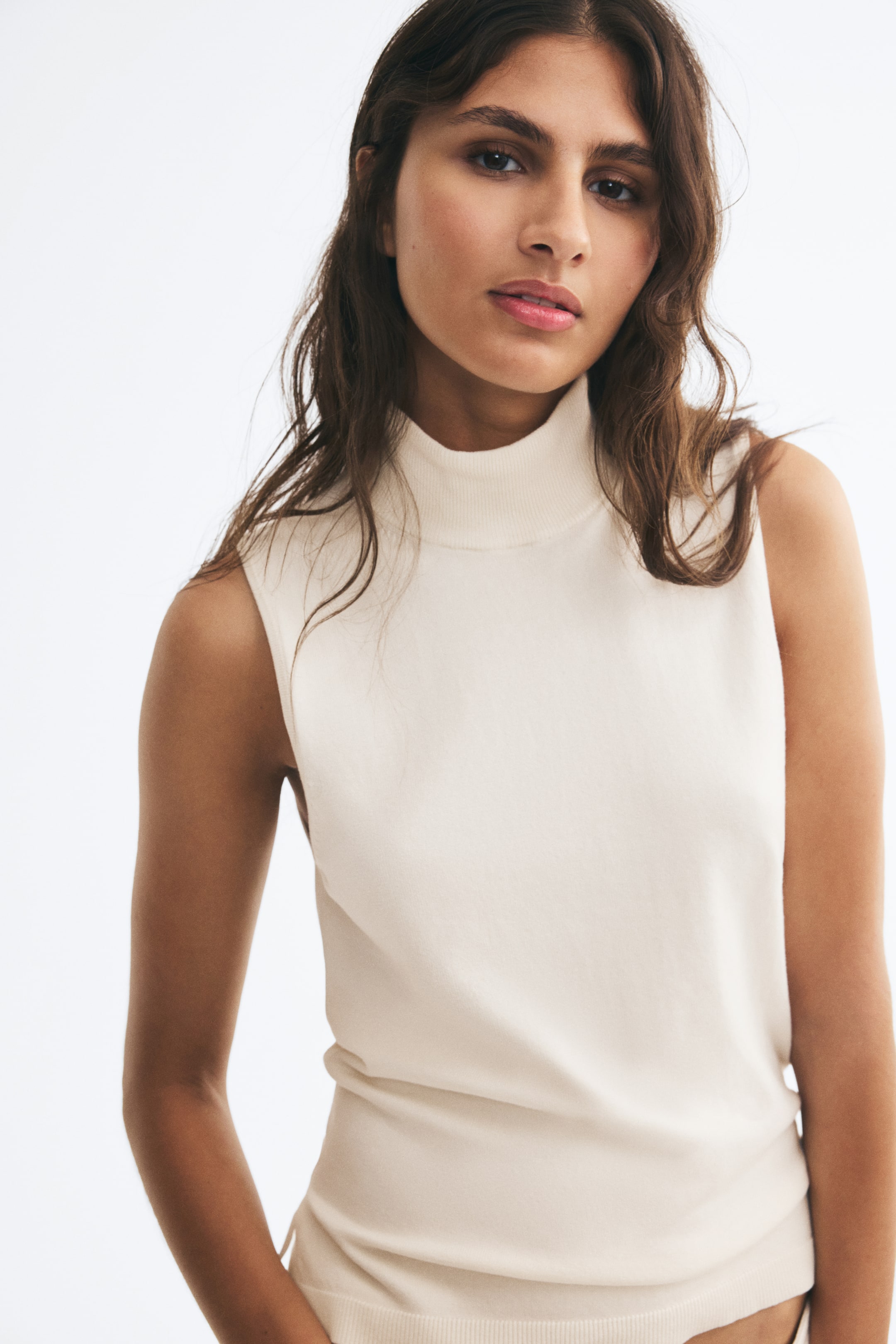 View larger image: The model is wearing a cream-colored, sleeveless knit vest. The fitted vest features a ribbed high neck and a ribbed hem.