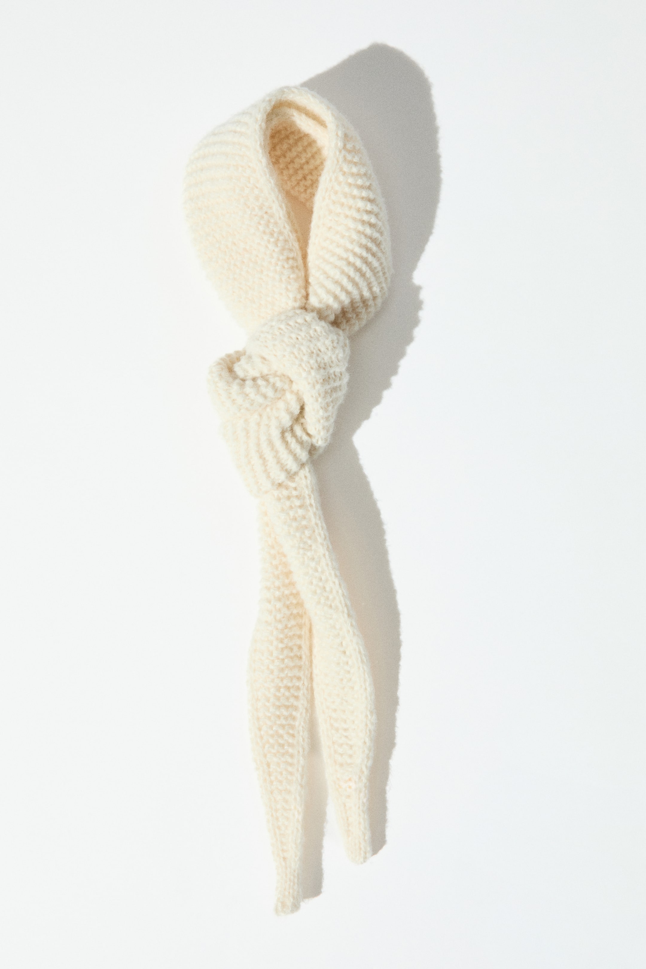View larger image: A cream-coloured scarf in a soft garter stitch knit is shown front-facing, tied in a loose knot that creates a loop at the top and two hanging tails. The scarf tapers down to distinct pointed ends, showcasing its textured, ribbed-like surface.
