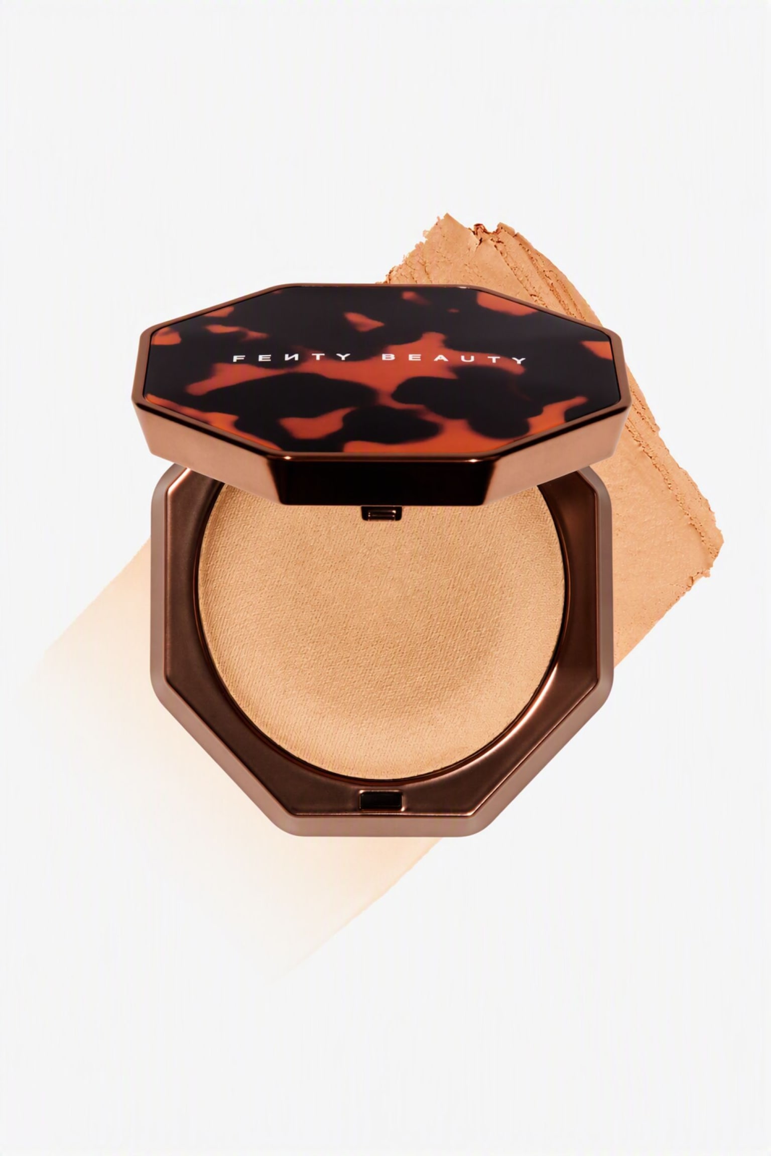 En Ã¶ppen, oktagonal bronzepuderask med lock i skÃ¶ldpaddsmÃ¶nster och varumÃ¤rkesnamnet "Fenty Beauty" visar en ljusbrun pressad bronzer. En diagonal, texturerad utstrykning av samma bronzerton strÃ¤cker sig bakom puderdosan fÃ¶r att visa dess lena textur. - 1