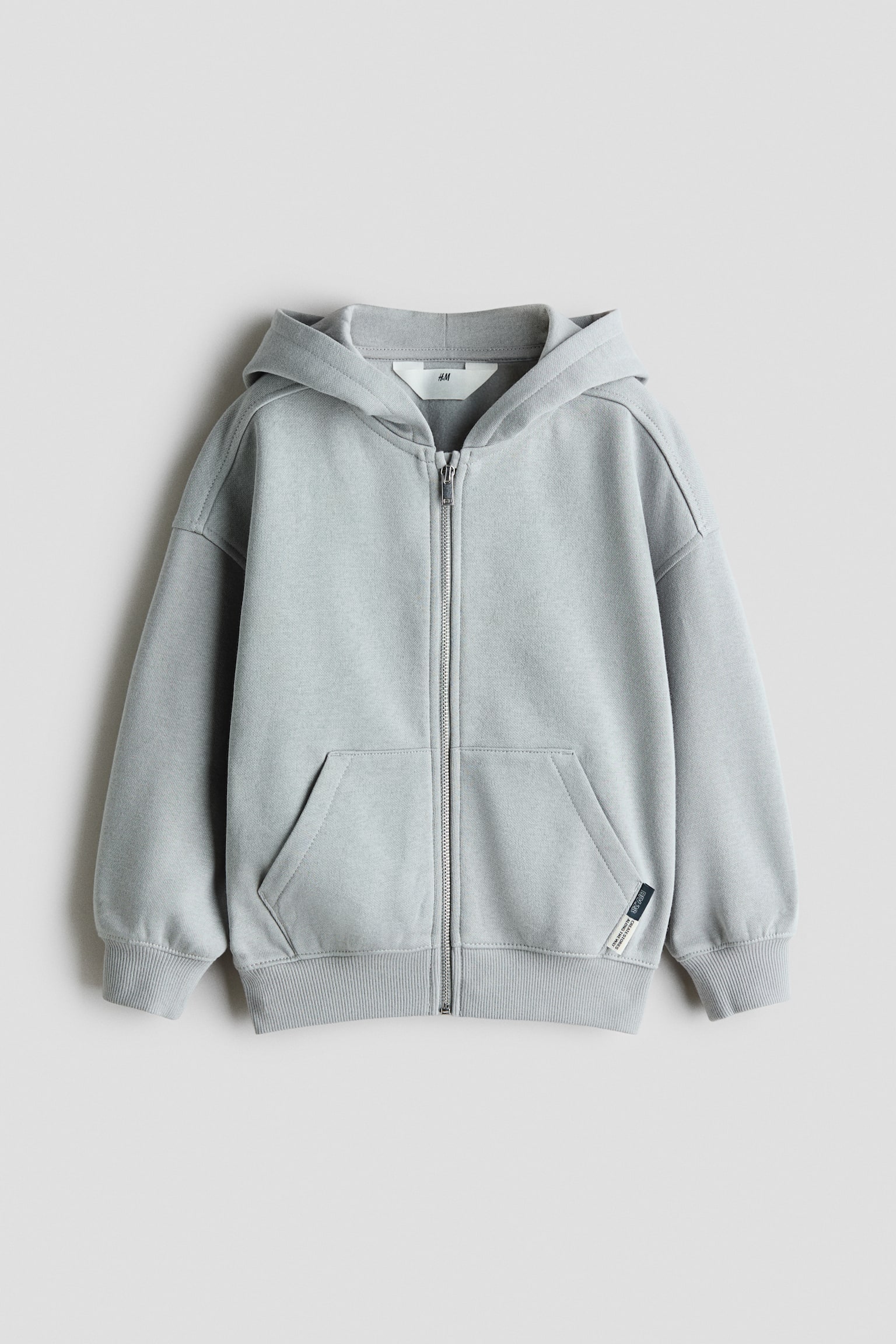 Un sweat Ã  capuche zippÃ© gris clair est prÃ©sentÃ© de face, mettant en valeur une fermeture Ã©clair mÃ©tallique sur toute la longueur, une capuche et des Ã©paules tombantes. Il prÃ©sente des poches kangourou fendues sur le bas de lâavant, des poignets cÃ´telÃ©s, un ourlet cÃ´telÃ© et une petite Ã©tiquette tissÃ©e sur la poche gauche.