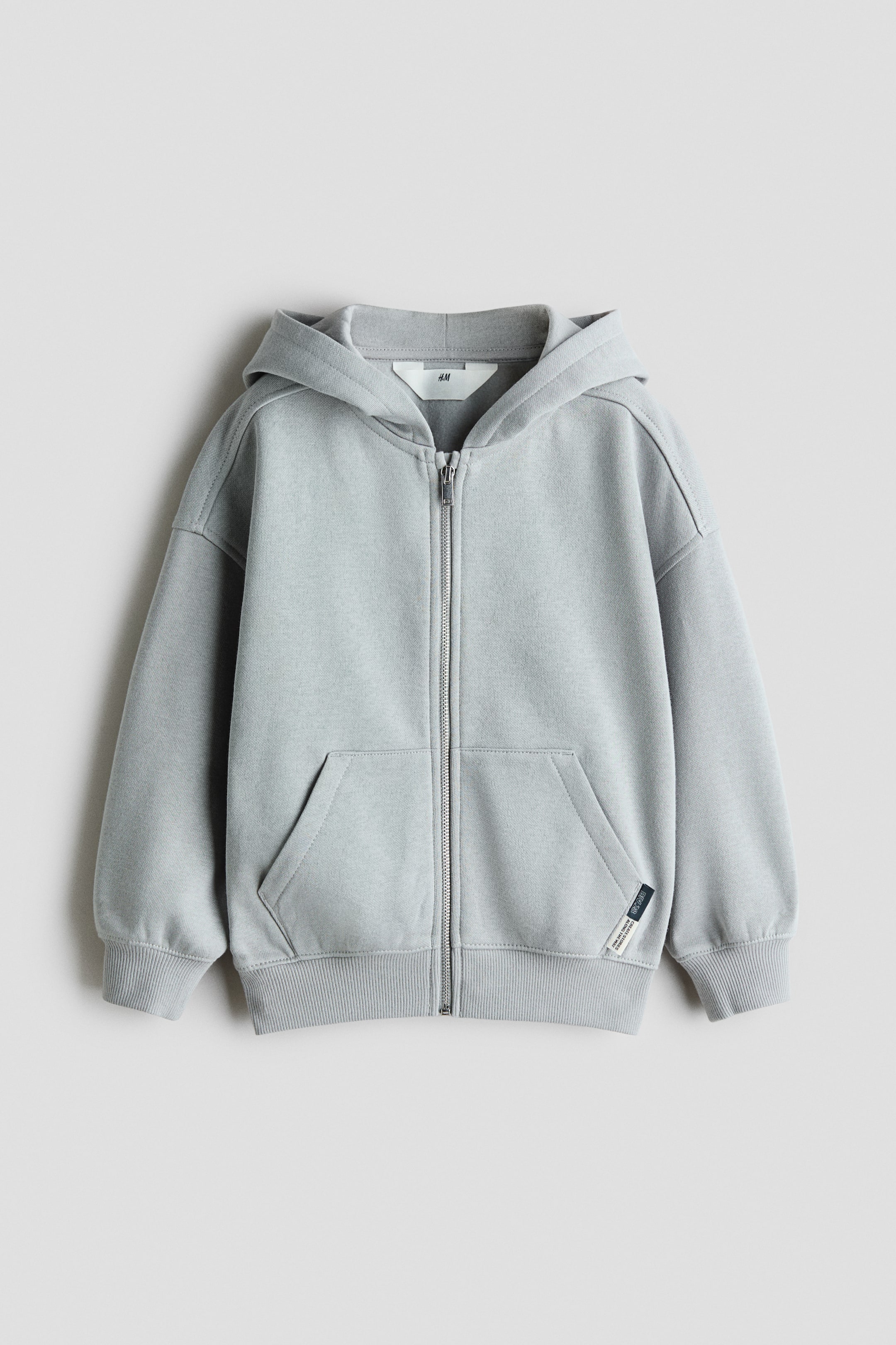 View larger image: A light grey zip-up hoodie is shown from the front, featuring a full-length metal zip, a hood, and dropped shoulders. It has split kangaroo pockets on the lower front, ribbed cuffs, a ribbed hem, and a small woven label on the left pocket.