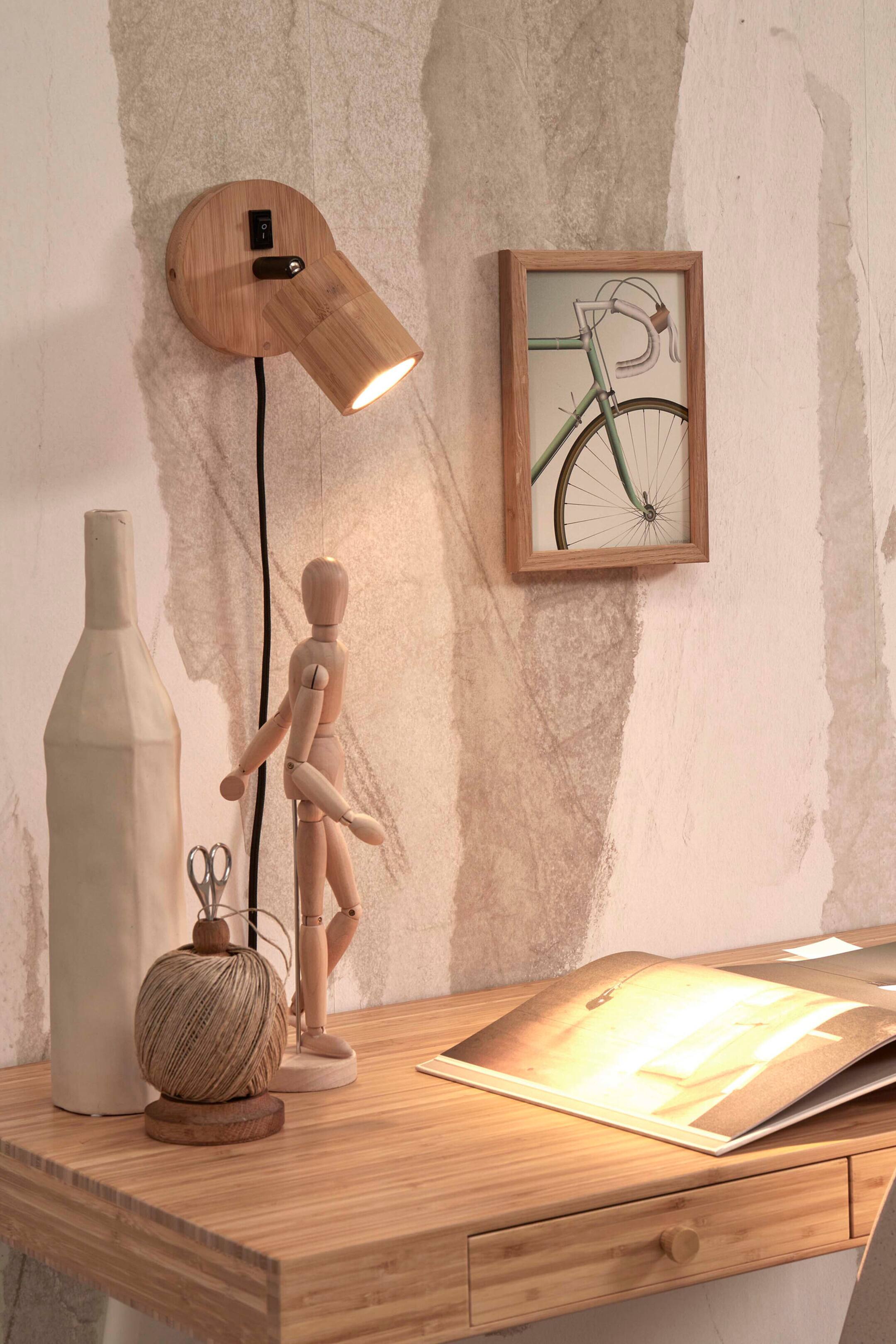 Agrandir l'image: Un spot en bois fixé au mur présente une base circulaire avec un interrupteur à bascule noir et un abat-jour cylindrique orientable. L’abat-jour est incliné vers le bas et diffuse une lueur chaleureuse, tandis qu’un cordon d’alimentation noir pend tout droit depuis la base.