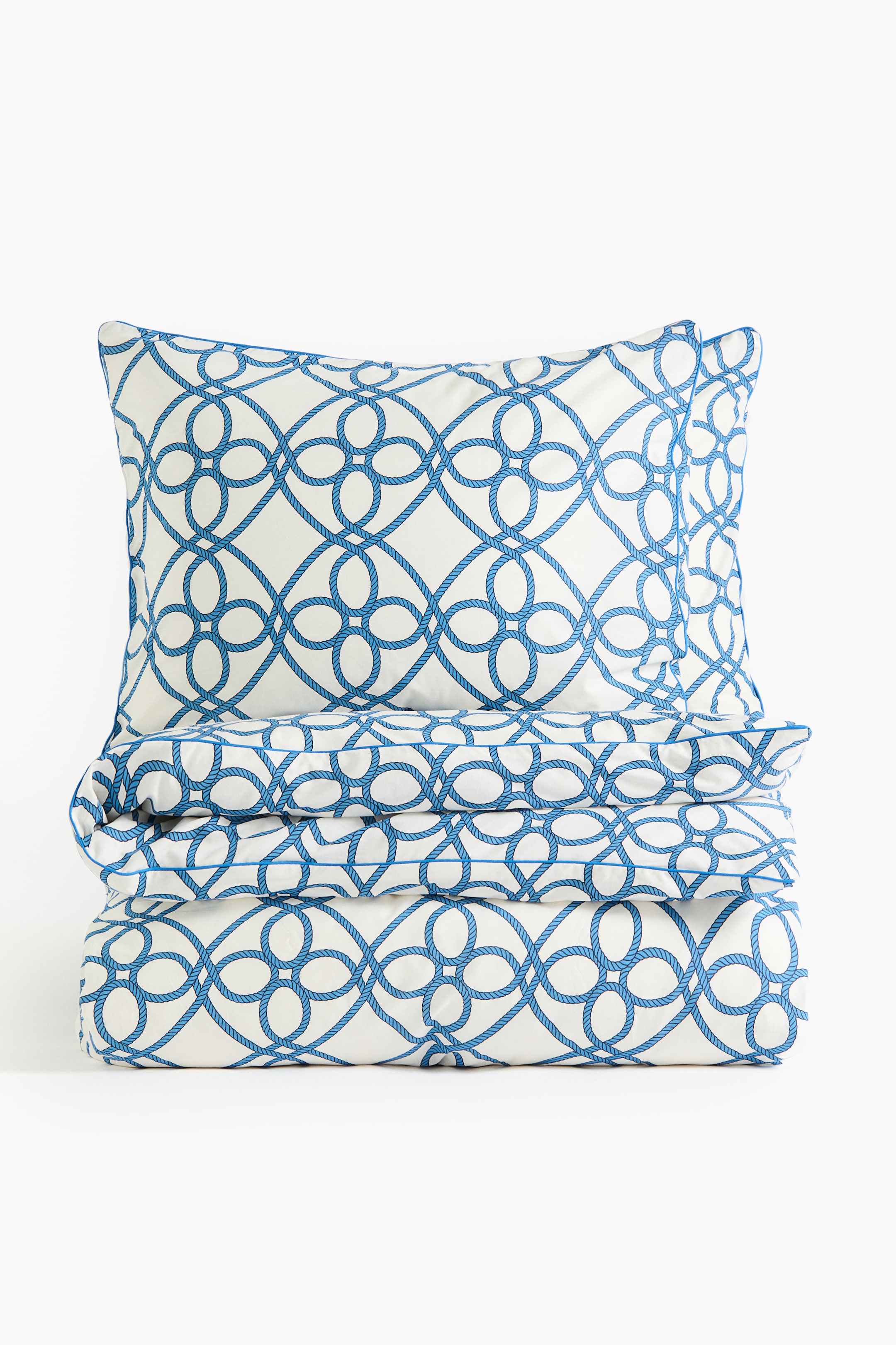 Ver imagen más grande: Un juego de ropa de cama presenta una tela blanca con un estampado repetido de cuerda azul que forma círculos geométricos entrelazados. Una almohada muestra su parte frontal y, debajo, hay un edredón doblado apilado; ambos muestran el estampado y un vivo azul en los bordes.