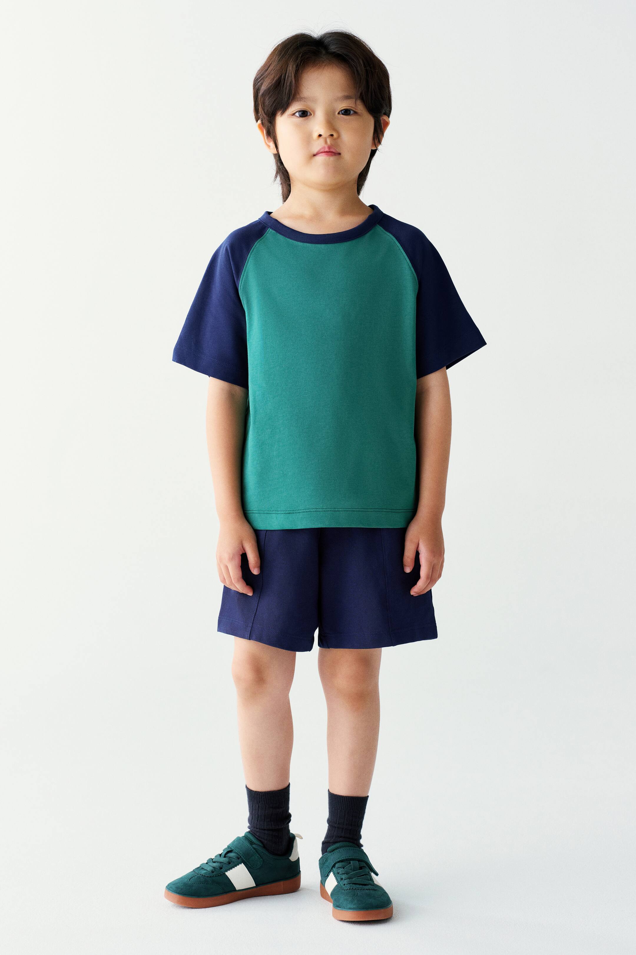 View larger image: The model is wearing a matching T-shirt and shorts set, featuring a green crewneck T-shirt with dark blue raglan sleeves paired with matching dark blue shorts. The outfit is styled with dark socks and green sneakers.