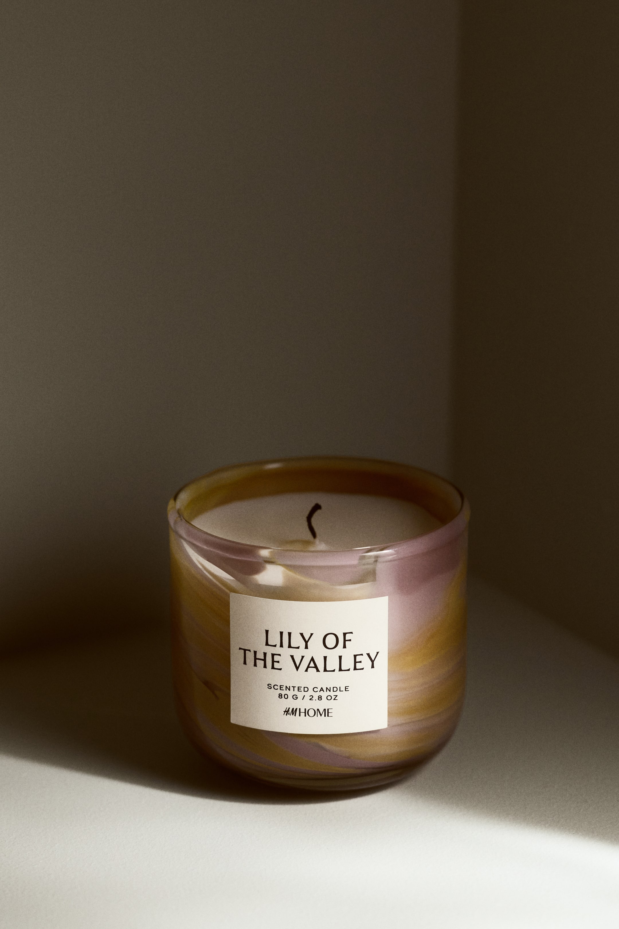 Ingrandisci l'immagine: Una candela profumata presenta un barattolo di vetro con motivi a spirale di tonalità viola e dorate. Un'etichetta bianca sul davanti mostra "LILY OF THE VALLEY," "CANDELA PROFUMATA," "80 G / 2.8 OZ," e "H&M HOME," mentre cera bianca riempie il barattolo con uno stoppino nero che emerge dal centro.