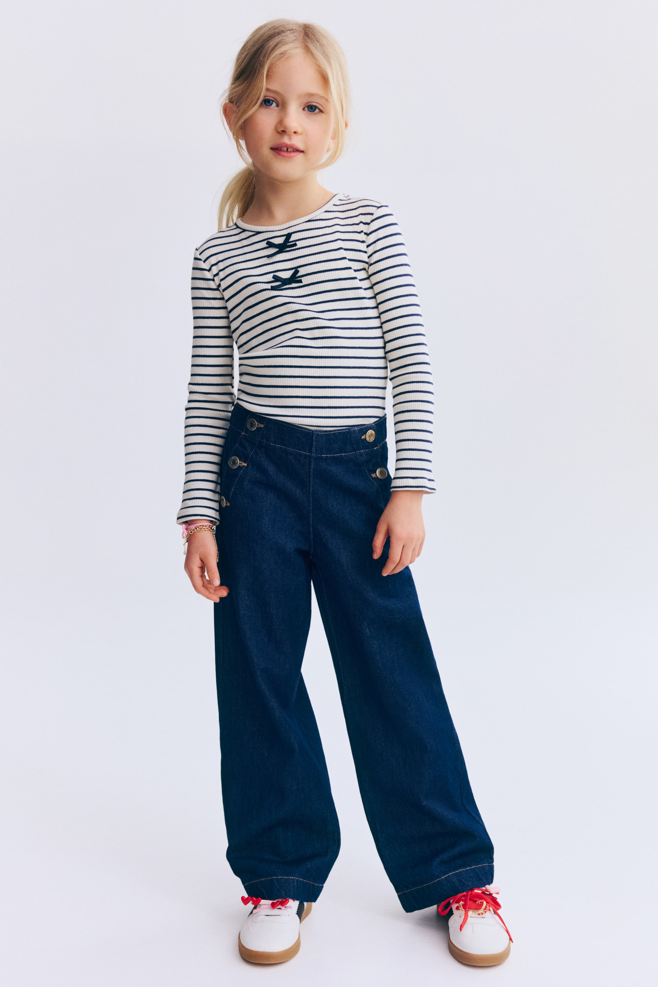 Agrandir l'image: Un top à manches longues côtelé présente des rayures horizontales blanches et bleu marine, rehaussé par deux petits nœuds bleu marine sur la poitrine. Il est porté rentré dans un pantalon en jean à jambe large de teinte foncée, présentant des boutons décoratifs dorés sur les côtés avant.