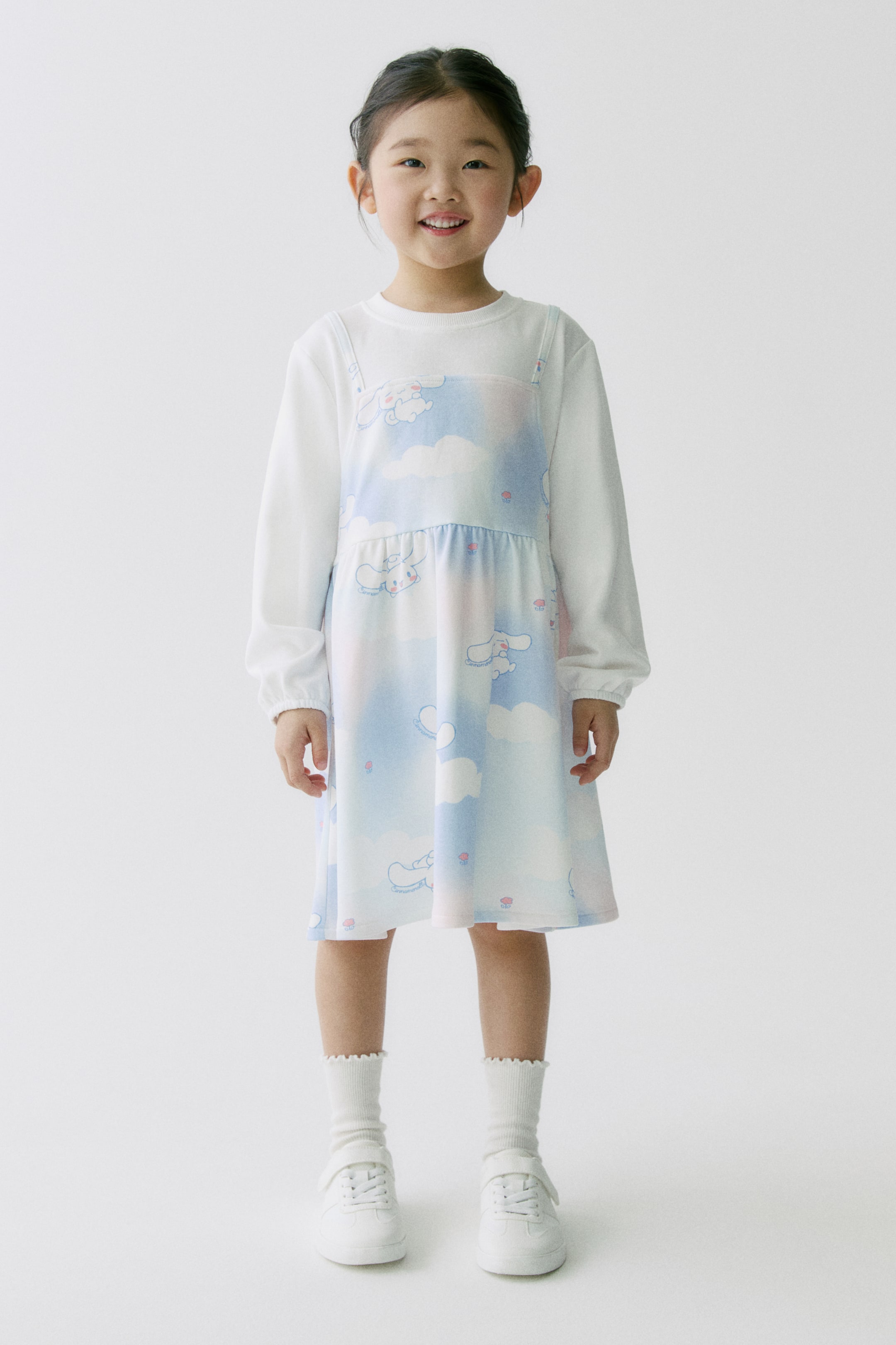 拡大画像を表示: ノースリーブのワンピースは、ライトブルー・ピンク・ホワイトのグラデーションに雲柄とカートゥーン風の動物柄が施され、白の長袖トップスに重ね着されています。ワンピースはスクエアネックで肩ひも付き、ウエストはギャザー入り、膝丈です。裾がスカラップの白いソックスと、白のベルクロストラップ付きスニーカーも合わせられています。