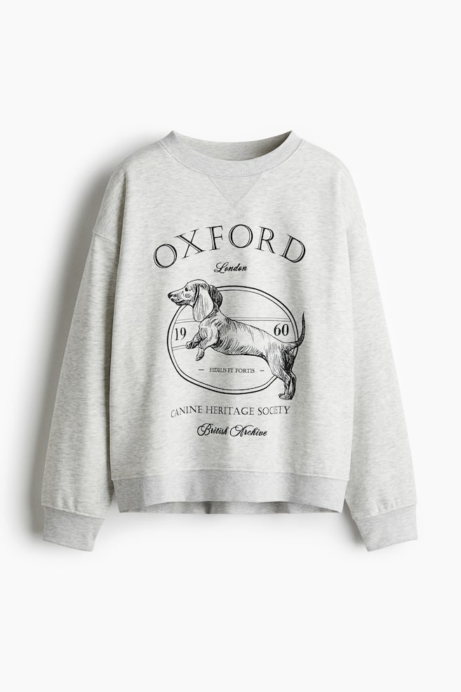 An oversized light gray heathered sweatshirt is presented FRONT, featuring a ribbed crew neck, dropped shoulders, long sleeves with ribbed cuffs, and a ribbed hem. A large black graphic motif, depicting a dachshund within a circular frame with text, is centered on the chest. - 1