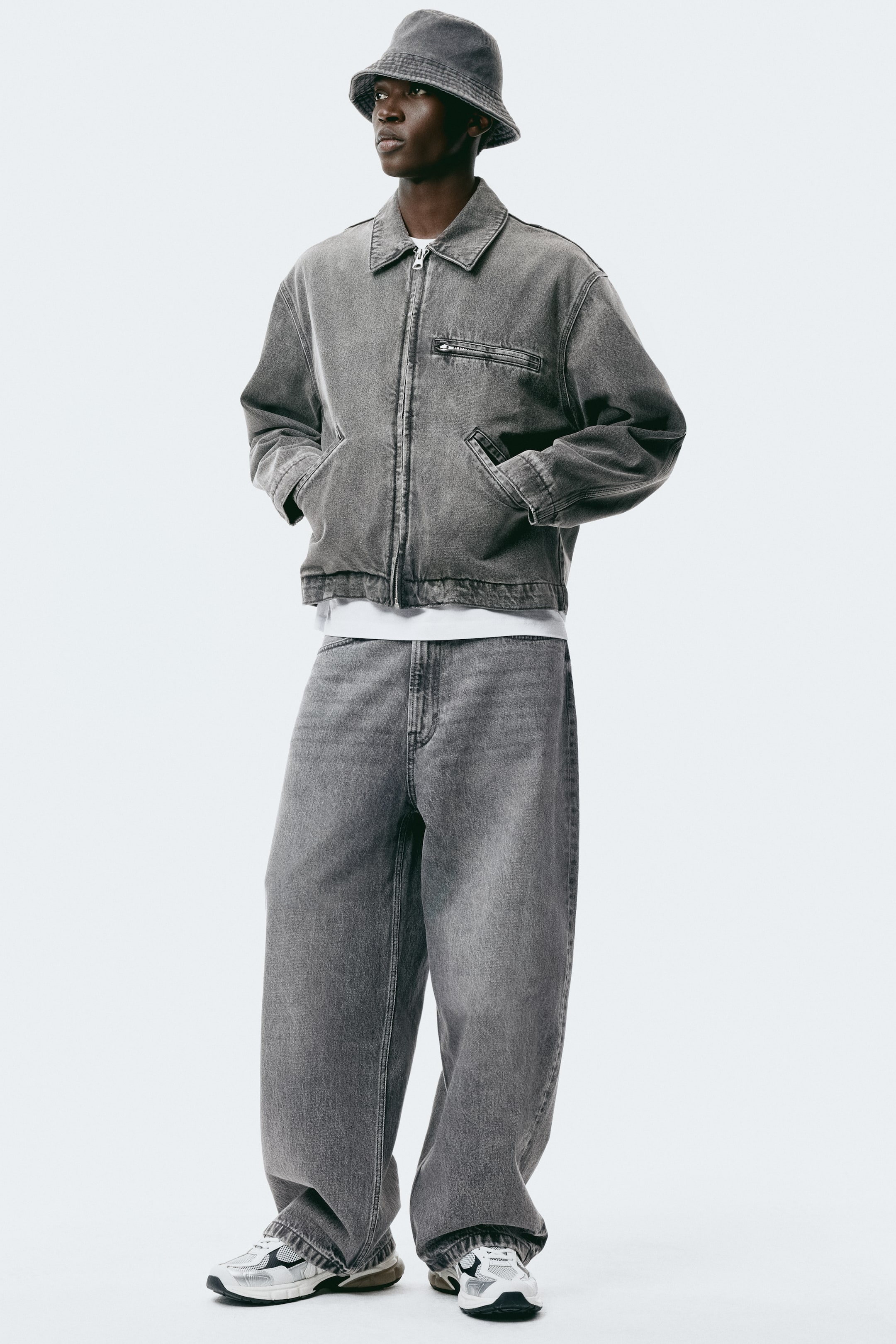 拡大画像を表示: グレーのウォッシュドデニムジャケットはジップ開閉のフロント、ポインテッドカラー、胸のジッパー付きポケット、そして2つのオープンウェルトポケットを備え、白いTシャツの上に重ねられています。マッチするグレーのワイドレッグジーンズはマルチトーンのスニーカーと合わせられ、同系色のグレーデニムバケットハットがコーディネートを完成させています。