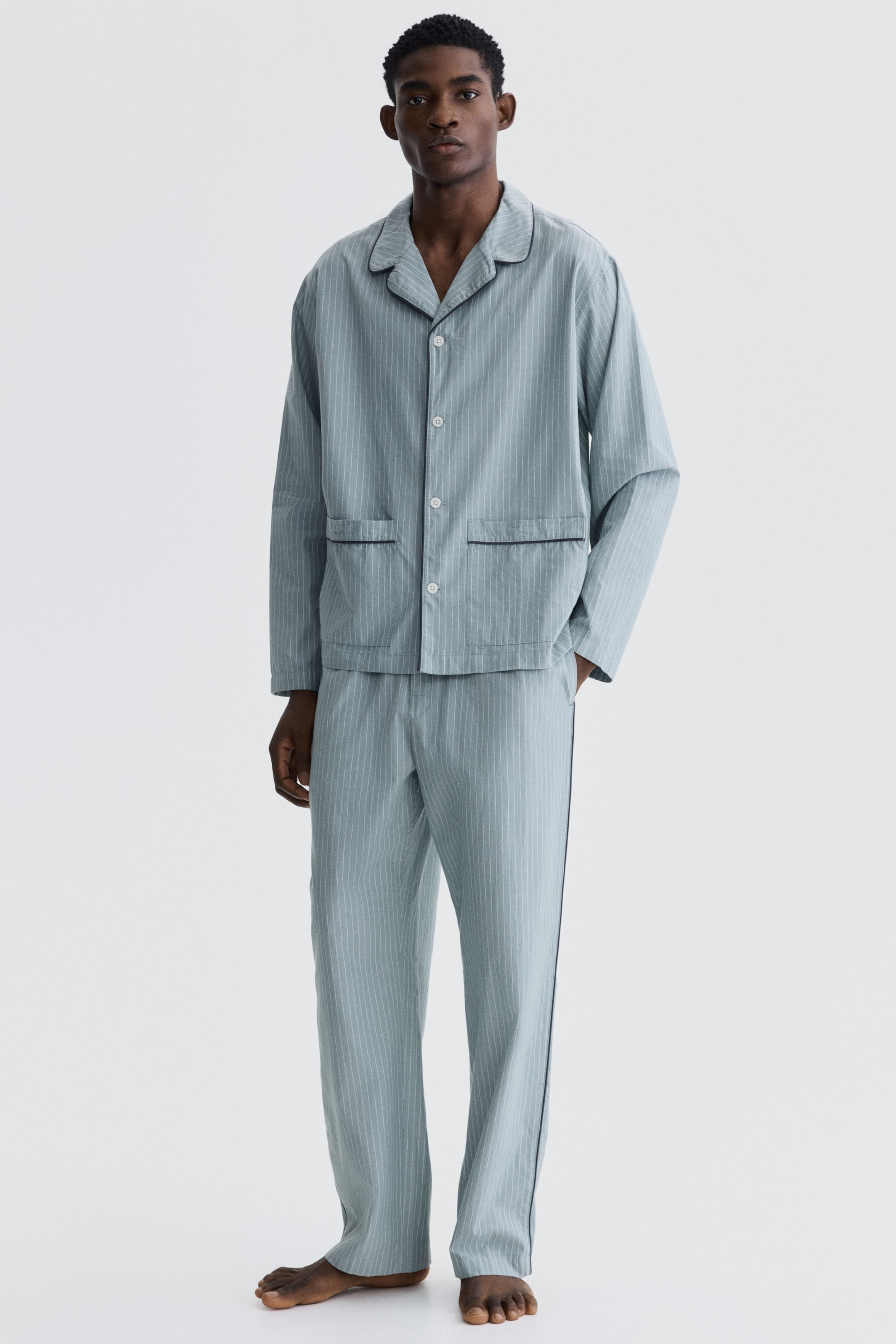 이미지 확대 보기: 연한 파란색 세로 줄무늬 파자마 세트는 노치 라펠이 있는 긴 소매 상의와 앞면에 흰색 버튼, 두 개의 앞쪽 패치 포켓이 있으며, 모든 부분은 어두운 파이핑으로 테두리가 있습니다. 일치하는 긴 바지도 세트를 완성하며, 옆 솔기에 어두운 파이핑이 특징입니다.