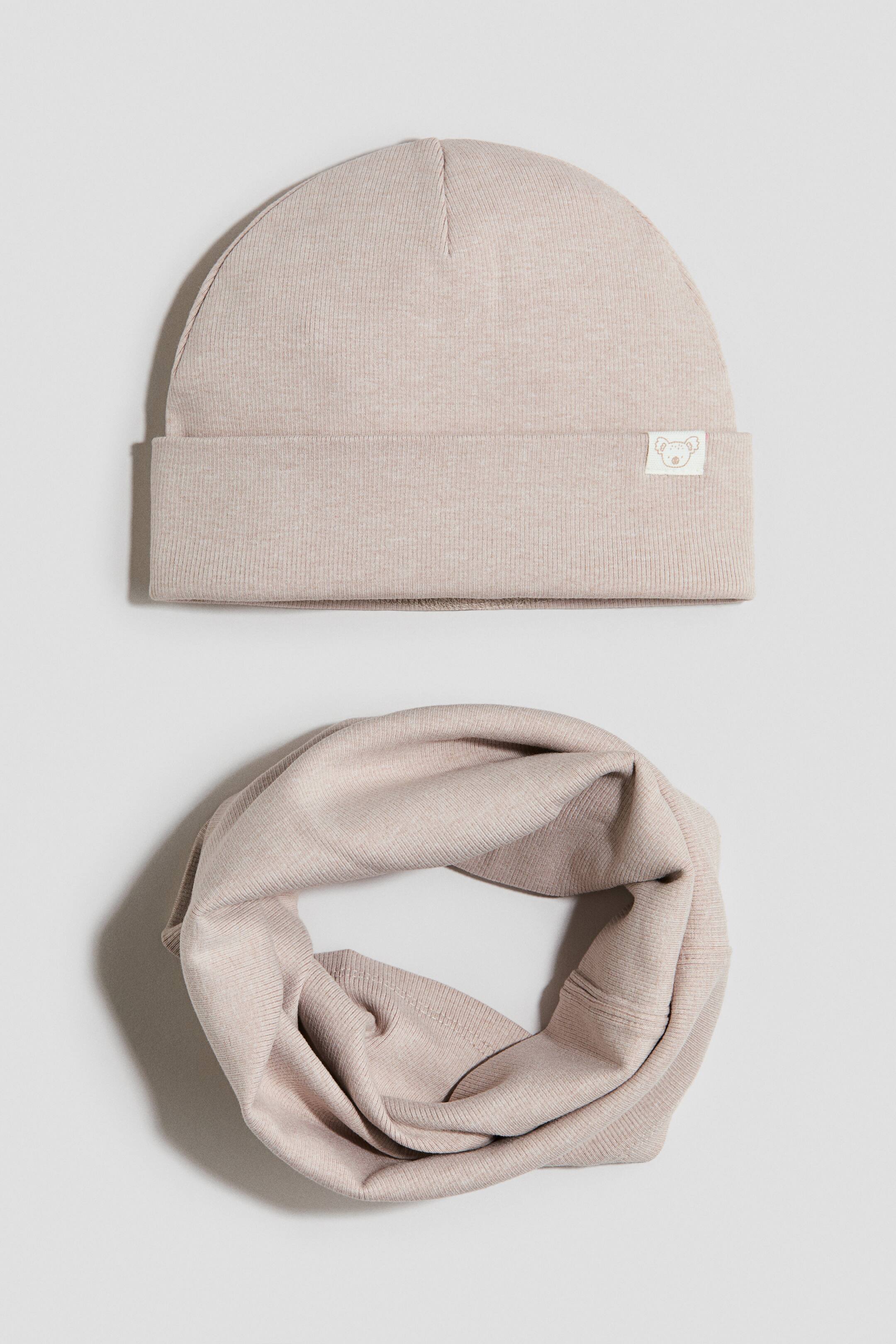 Grotere afbeelding bekijken: Een tweedelige lichtbeige jerseyset toont een beanie in vooraanzicht met een omgeslagen boord en een klein label met de contour van een beer. Een bijpassende nekwarmer is in een cirkel gevouwen en toont de geribde jerseystructuur.