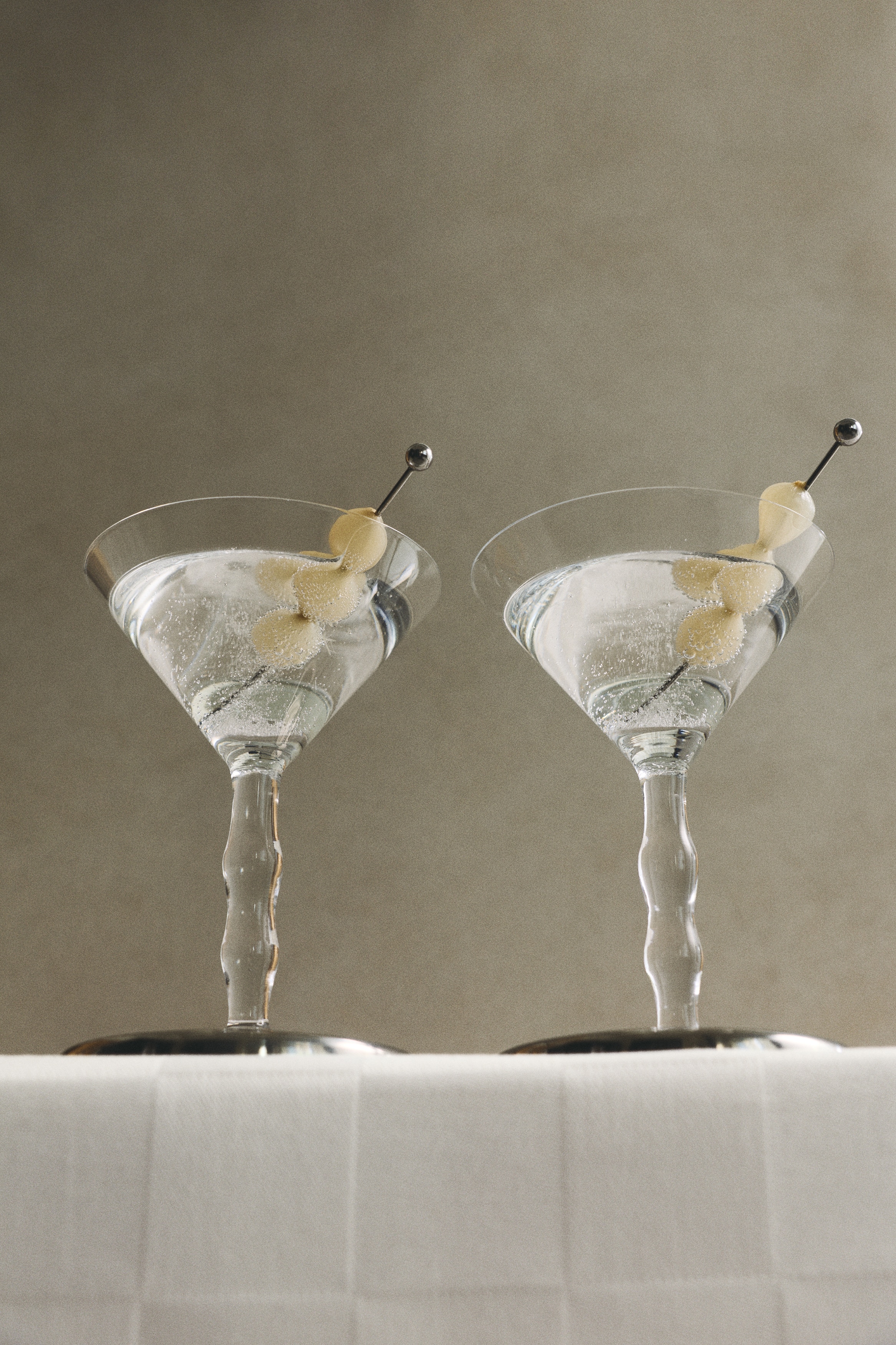 Une paire de verres à martini en verre transparent est disposée côte à côte, tournée vers lâavant, avec des coupes coniques, des pieds ondulés sculptés et des bases circulaires de ton argenté. Chaque verre contient un fin pic à cocktail en métal argenté avec une petite extrémité sphérique, posé en diagonale à lâintérieur de la coupe.