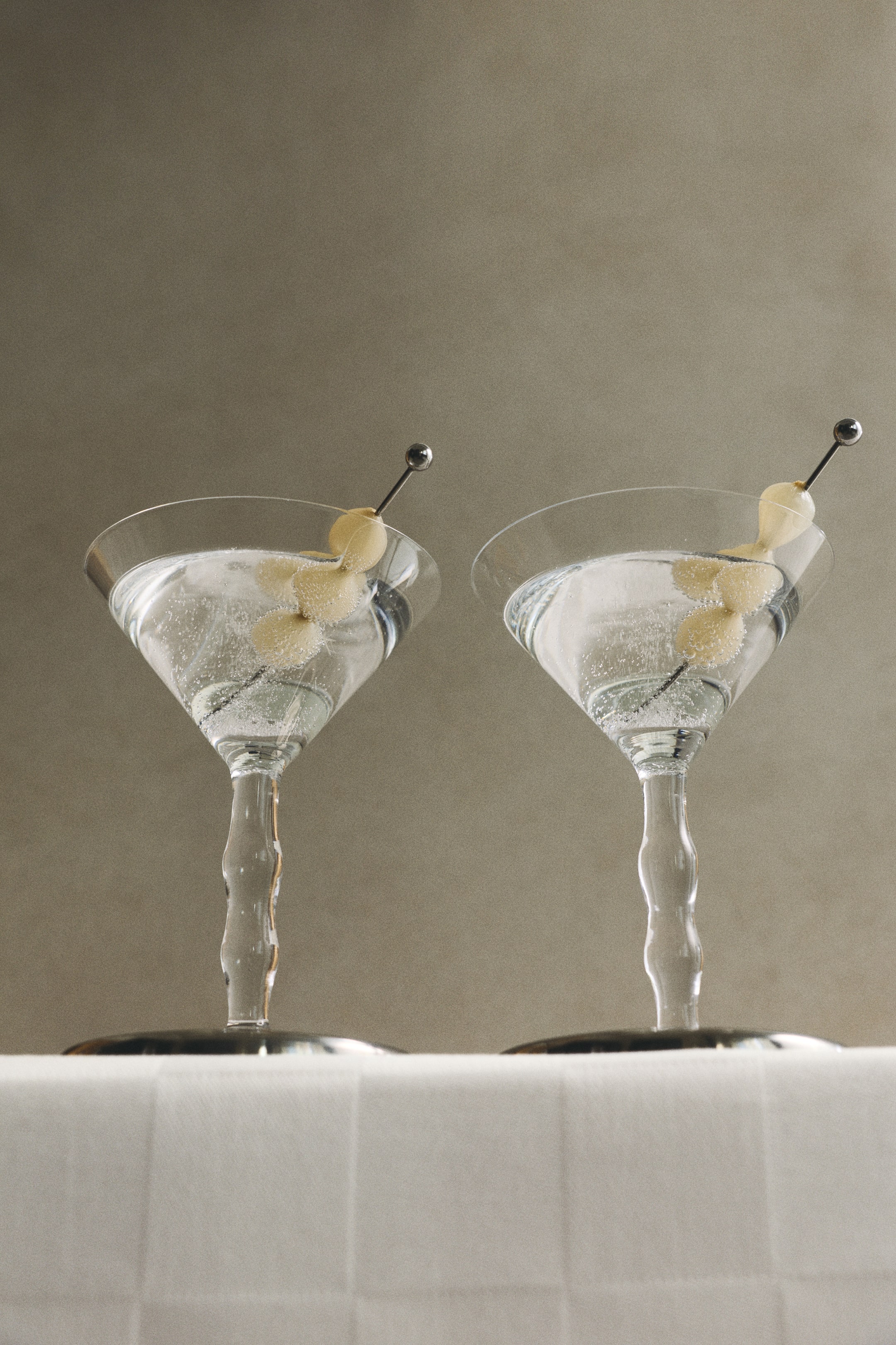 Ver imagem maior: Dois copos de cocktail em vidro transparente são apresentados de frente, com hastes altas e onduladas distintivas, assentes em bases circulares de aço inoxidável. Cada copo contém uma bebida transparente, guarnecida com um palito de cocktail em aço inoxidável a segurar três cebolas de cocktail.