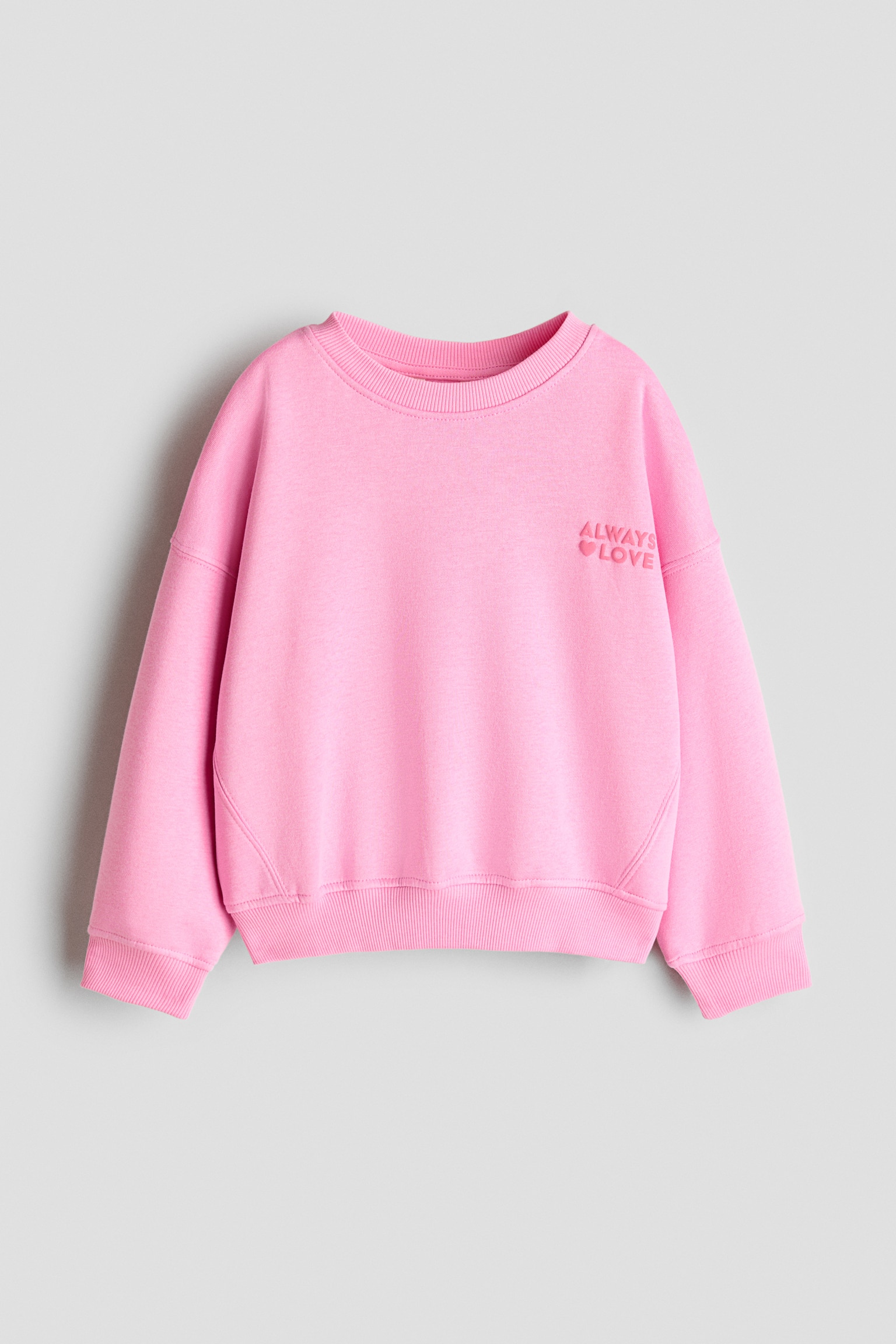 A pink sweatshirt is shown from the front, featuring a relaxed fit with dropped shoulders, a crew neckline, ribbed cuffs, and a ribbed hem. The sweatshirt has a small tonal embroidered text on the left chest that reads "ALWAYS" above "LOVE" with a heart icon.