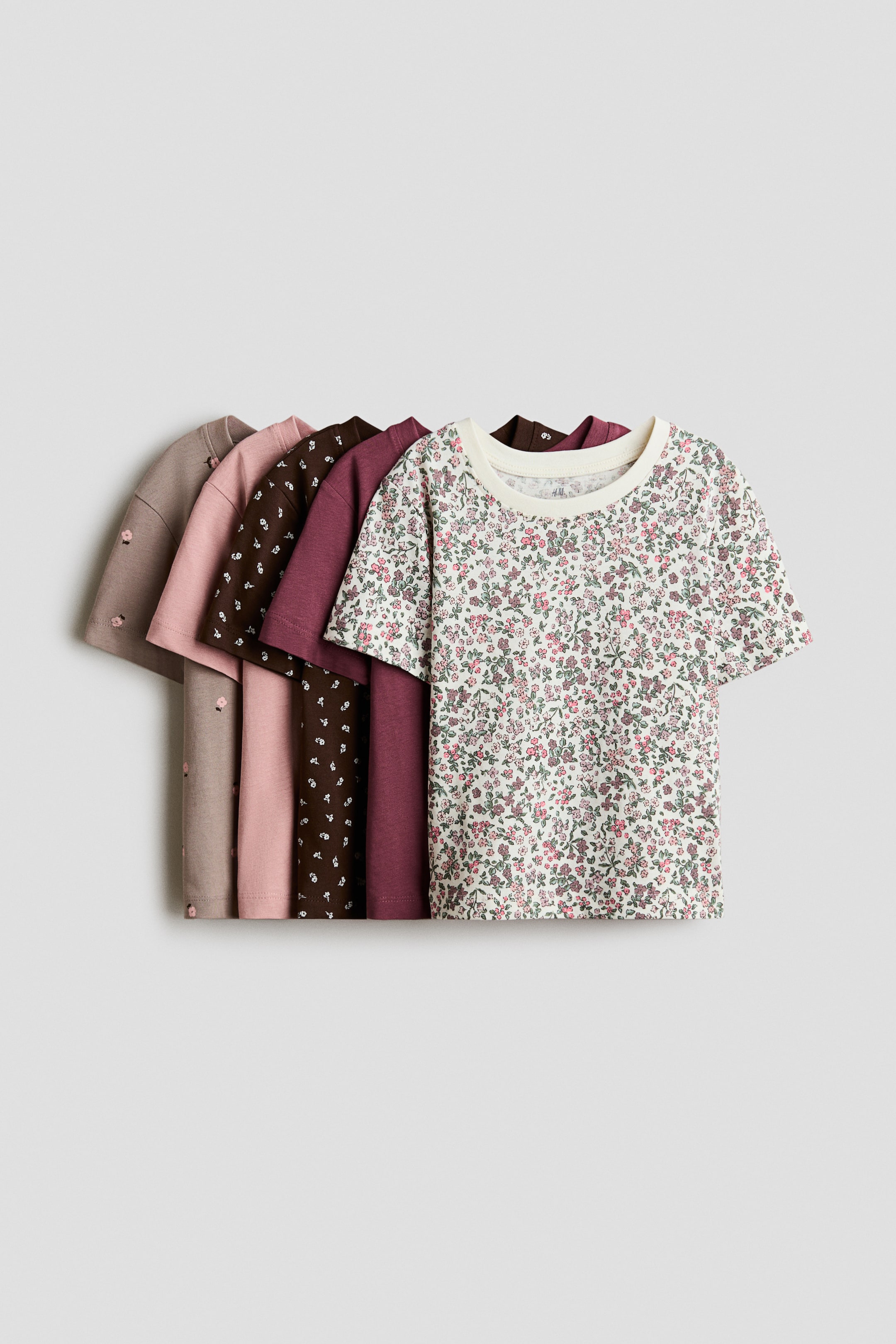View larger image: Five short-sleeved cotton jersey T-shirts are layered and facing forward to showcase their distinct designs. The front T-shirt is cream-colored with an all-over pink and green floral pattern, while the shirts behind it include solid plum and dusty pink, as well as patterned styles in dark brown and taupe.