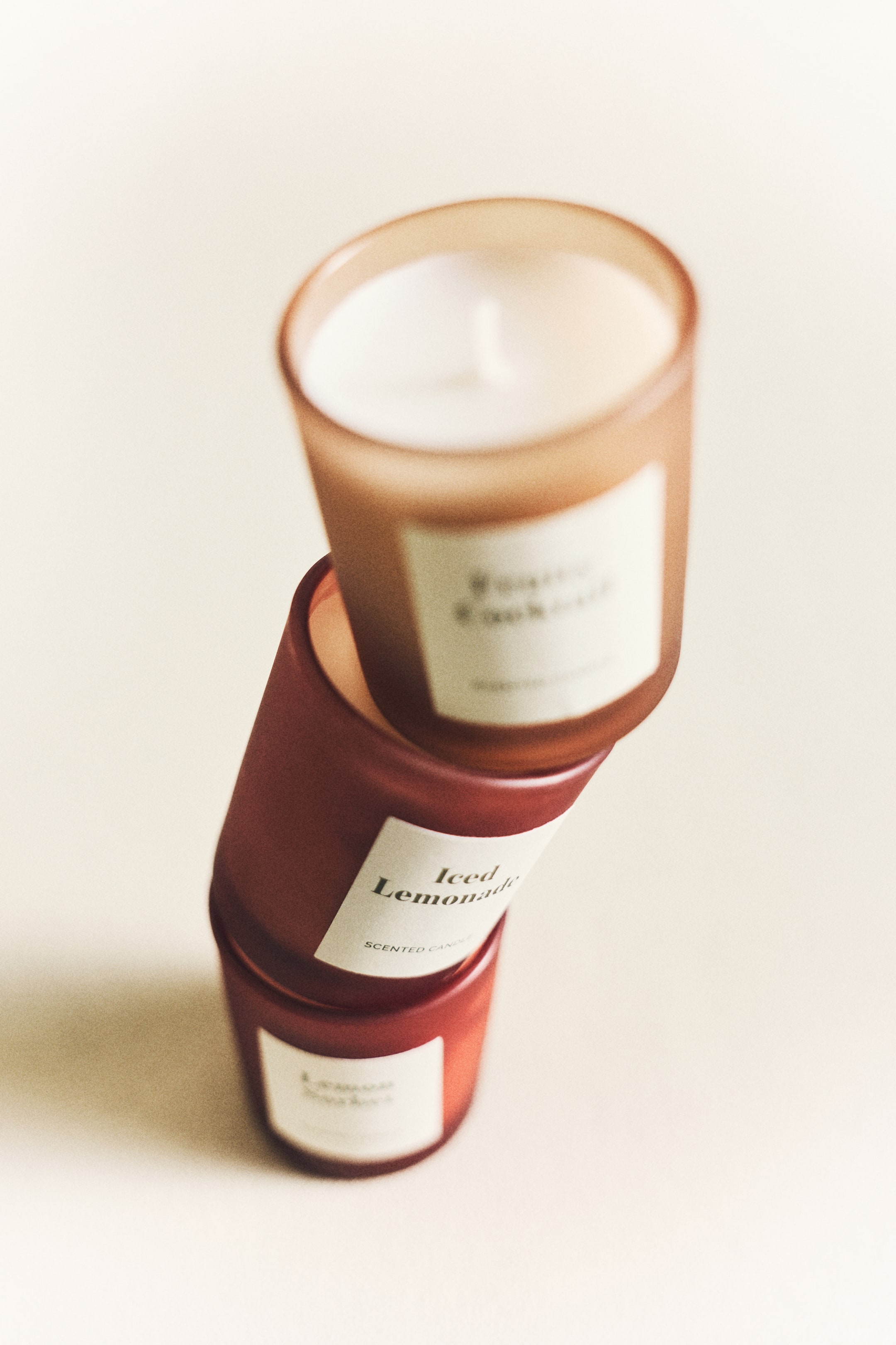 View larger image: Three scented candles are stacked vertically, each nestled in a glass holder. The top candle is housed in a translucent amber holder, while the two lower candles sit in reddish-brown holders. Each candle features a white label with black text, including 'Iced Lemonade' on the middle one.