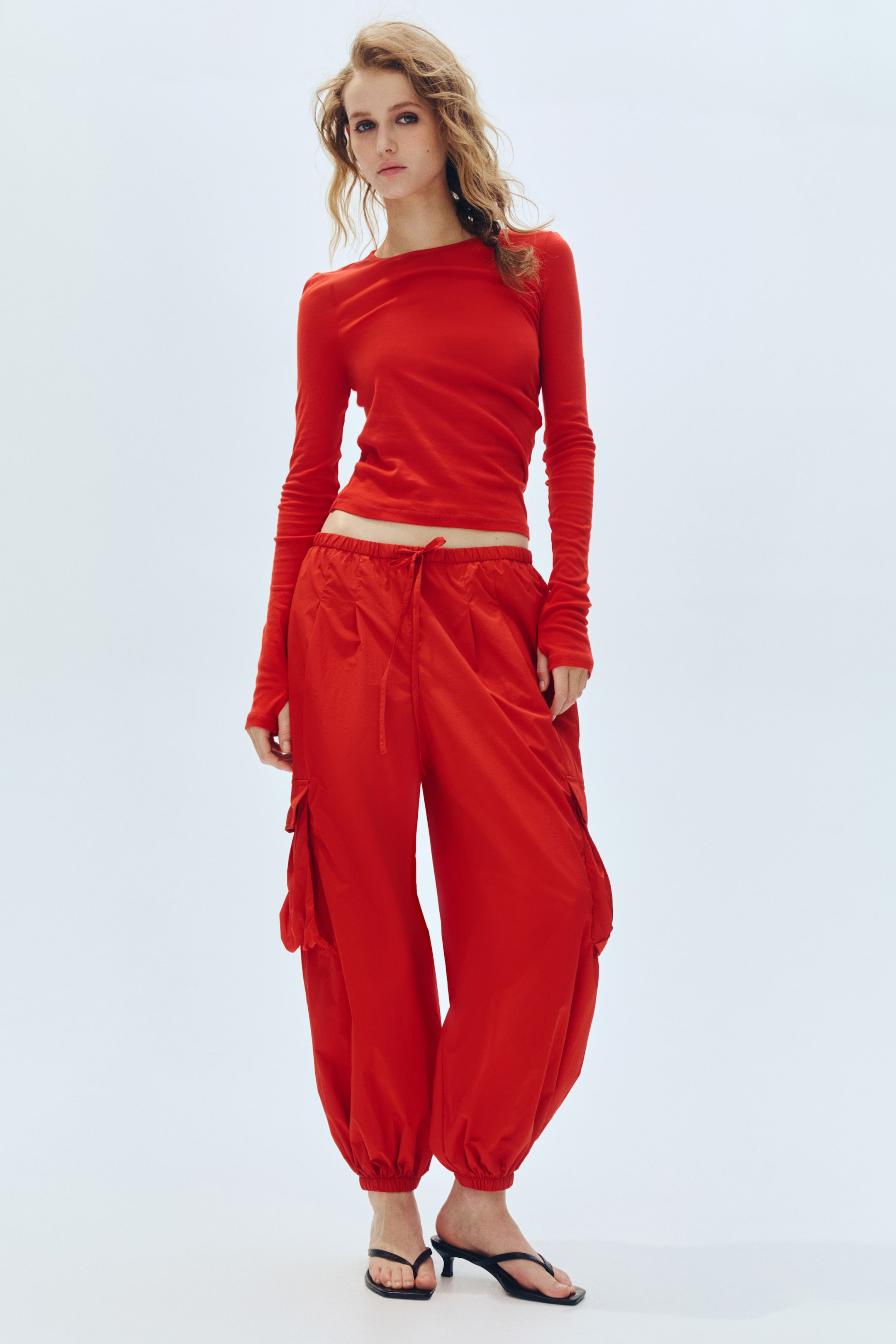 View larger image: Presenting a red long-sleeve, fitted crew neck top featuring extra-long sleeves with thumbhole details, paired with red wide-leg cargo pants. The pants showcase a drawstring waist, side cargo pockets, and gathered ankles, while the top is styled to crop just above the waist.